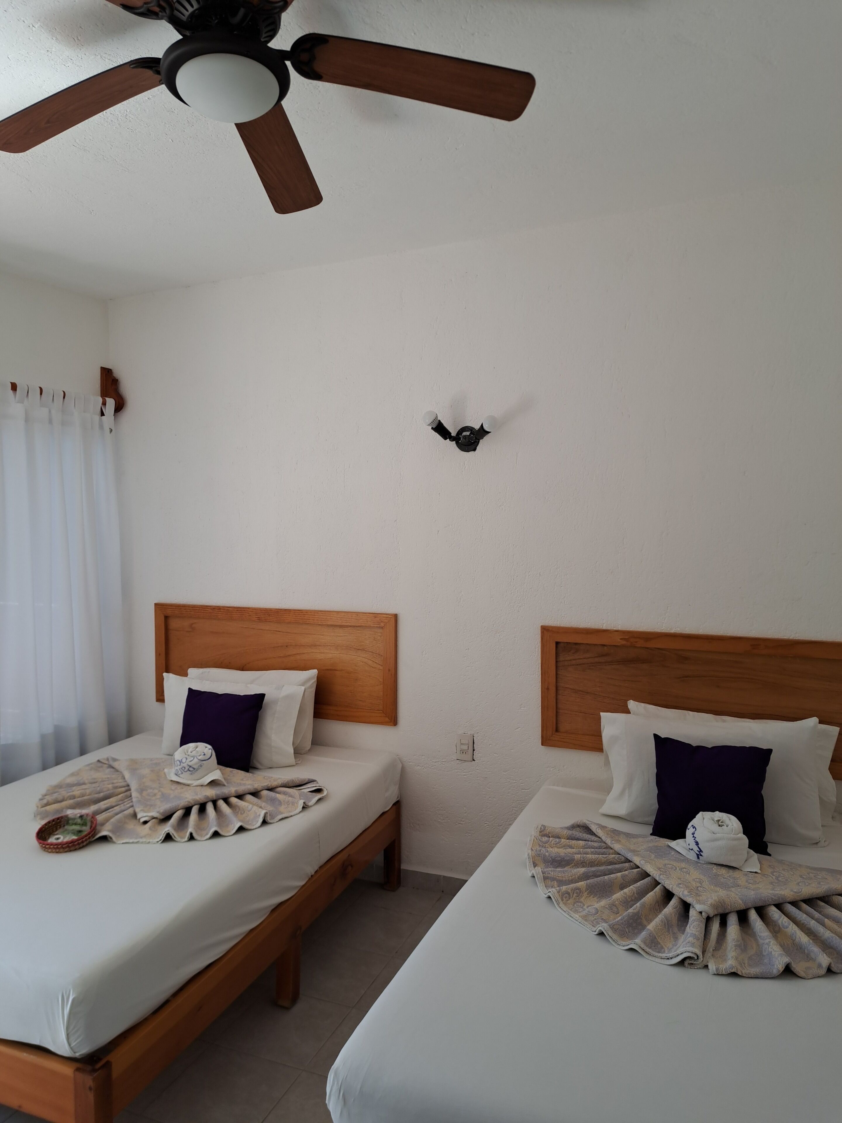 Une chambre d’hôtel avec deux lits en bois, du linge de lit blanc et un ventilateur de plafond.