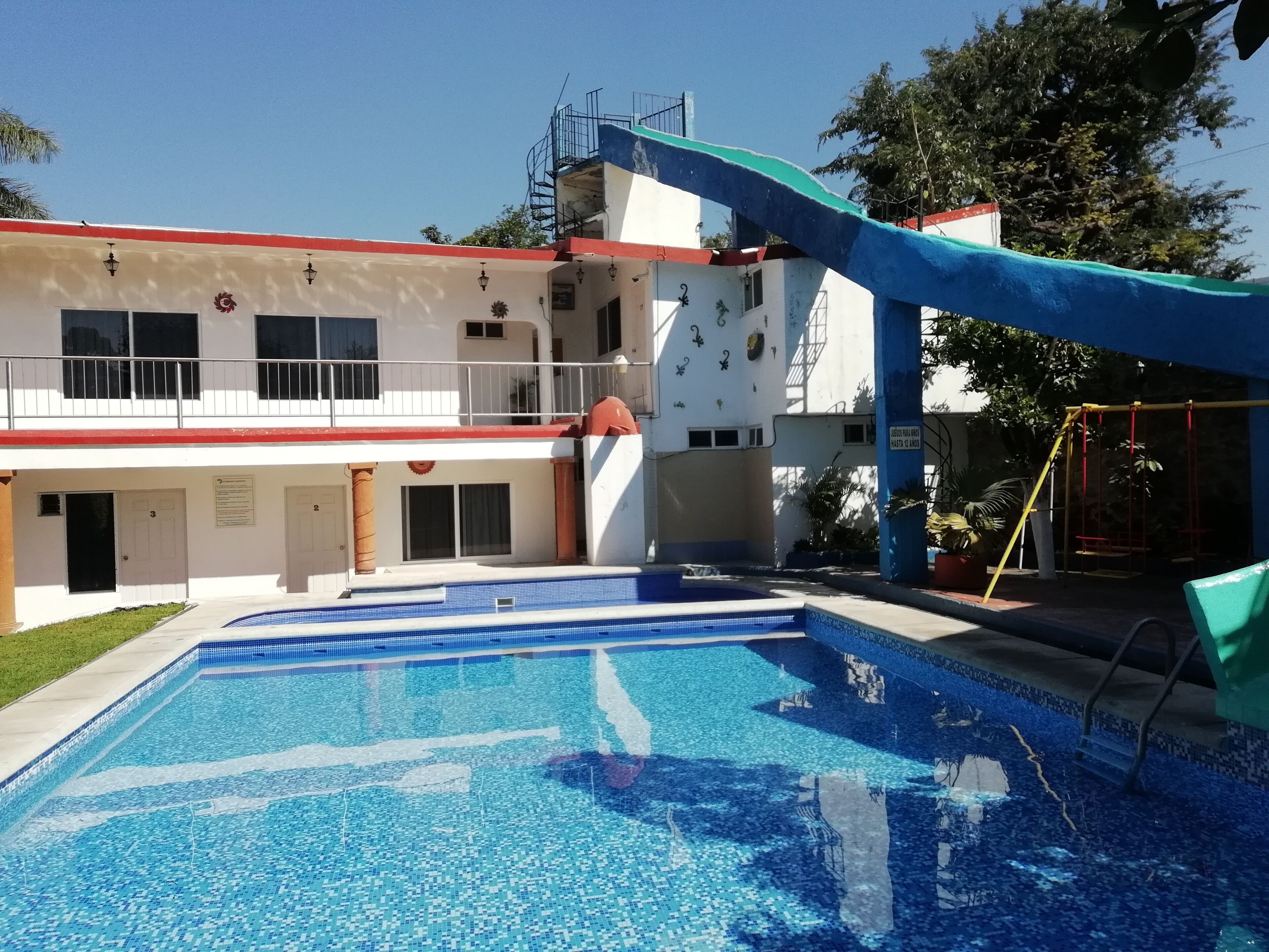 A swimming pool with a slide, a building with multiple windows and balconies, and a playground area with swings and climbing structures.