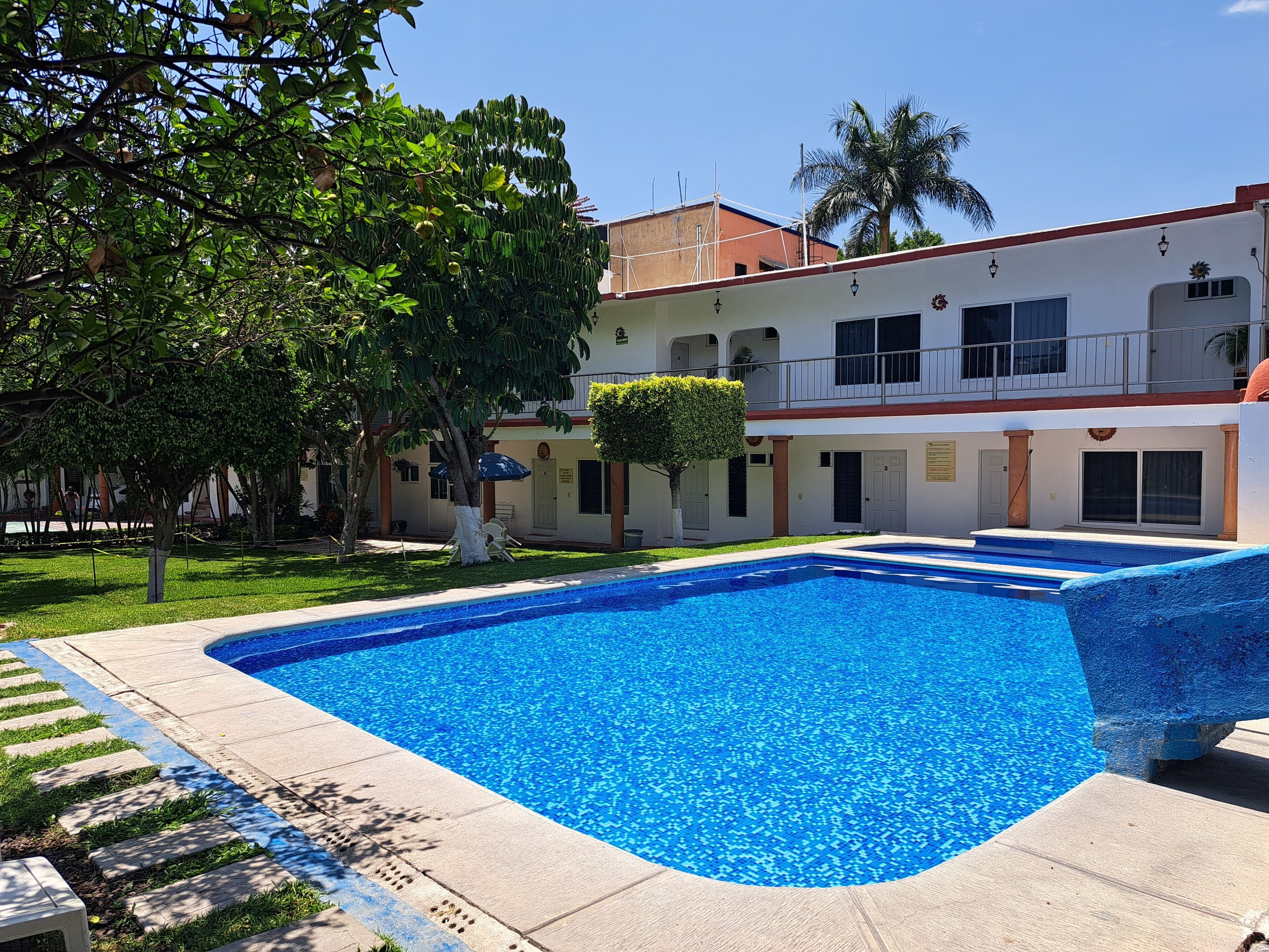 Una piscina con agua azul cristalina, rodeada por un solárium con baldosas. Se ven árboles y un edificio con balcón al fondo.