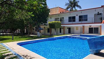 Una piscina con agua azul cristalina, rodeada por un solárium con baldosas. Se ven árboles y un edificio con balcón al fondo.