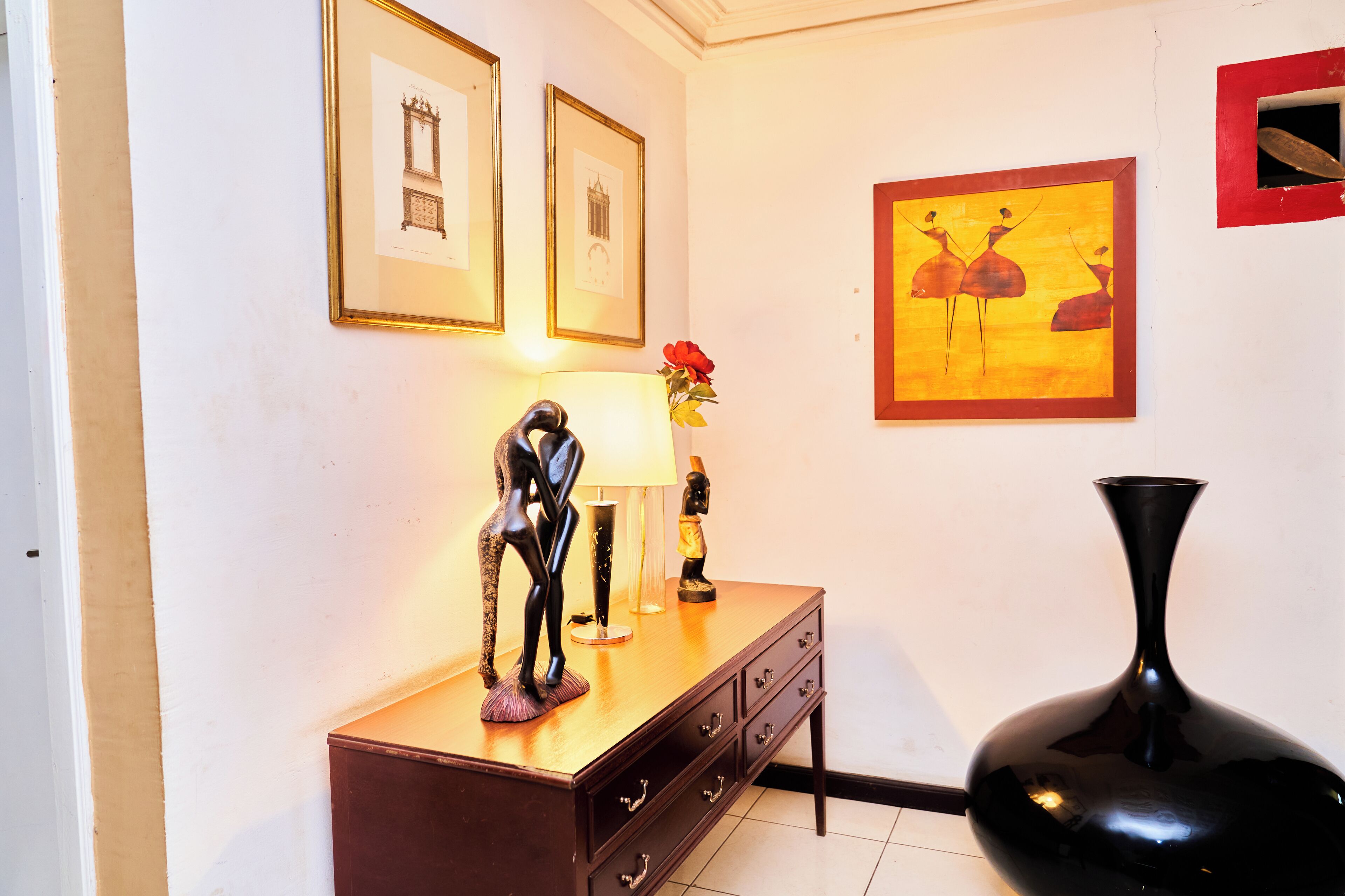 Een kamer met een houten dressoir, een zwarte vaas en kunstwerken in lijsten aan de muur.