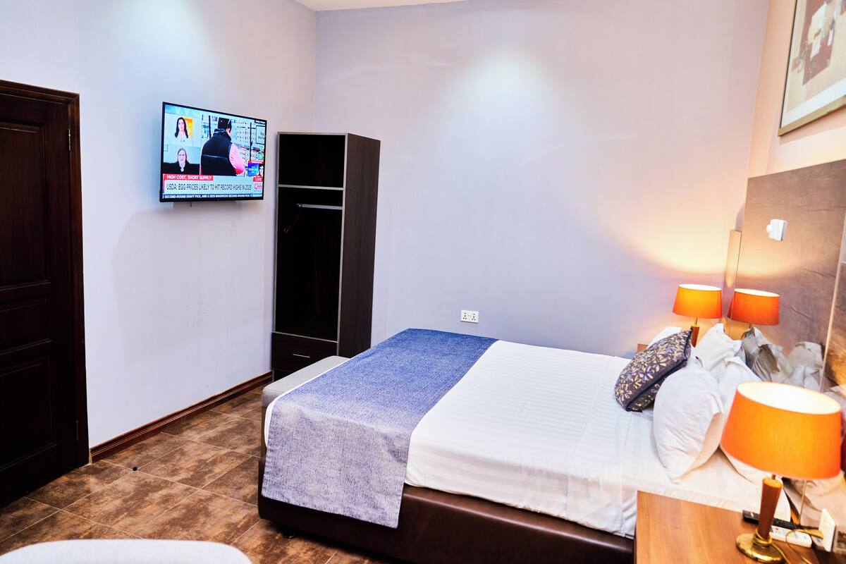 A hotel room with a bed, a TV on the wall, a wardrobe, and a nightstand with a lamp.