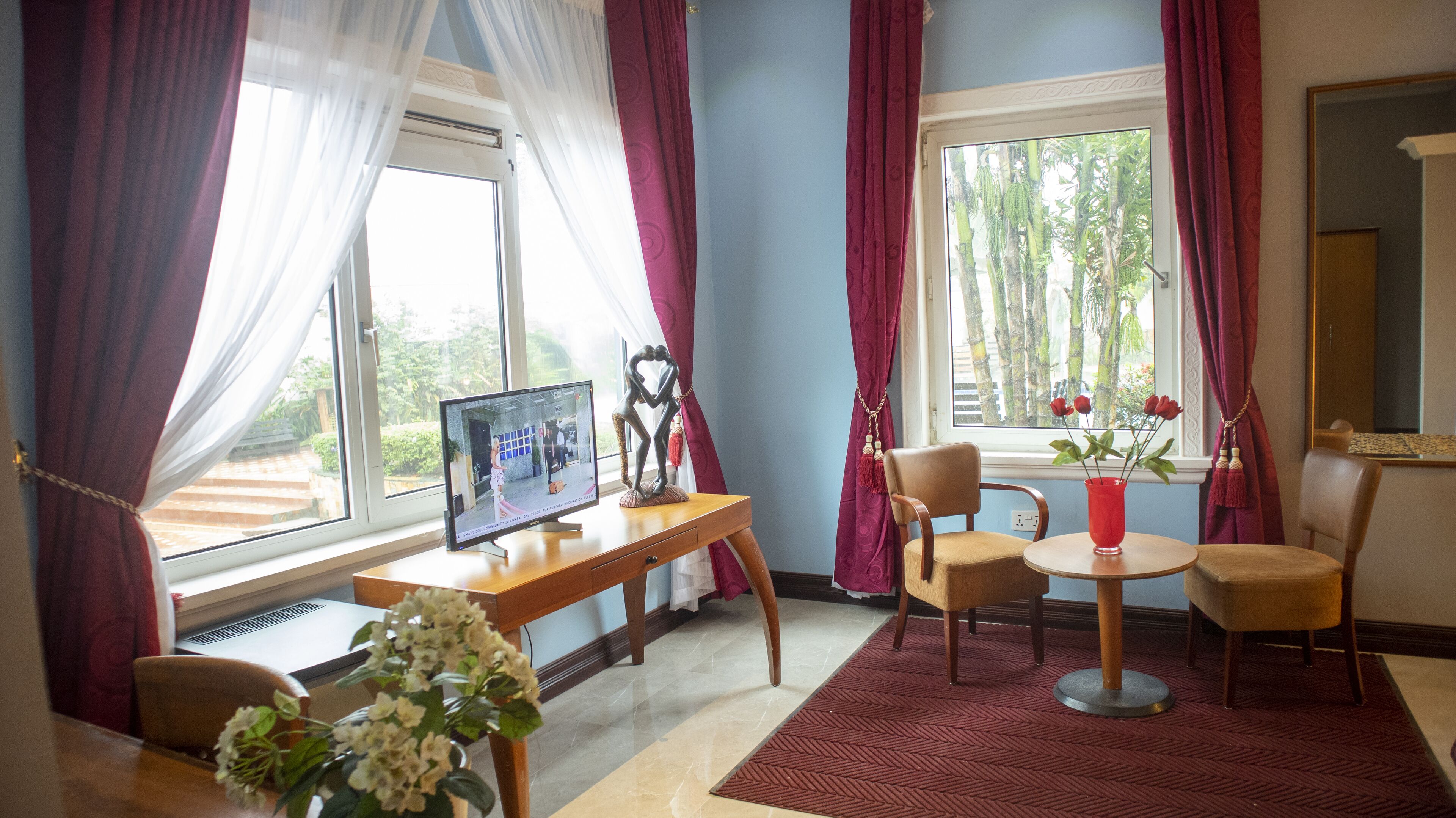 A room with a desk, computer, chairs, a small table with a vase, and a window with curtains.