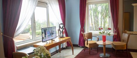 A room with a desk, computer, chairs, a small table with a vase, and a window with curtains.