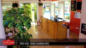 A front desk area with a wooden counter, a reception desk, and a large potted plant. - Grand Casamora (Accra)