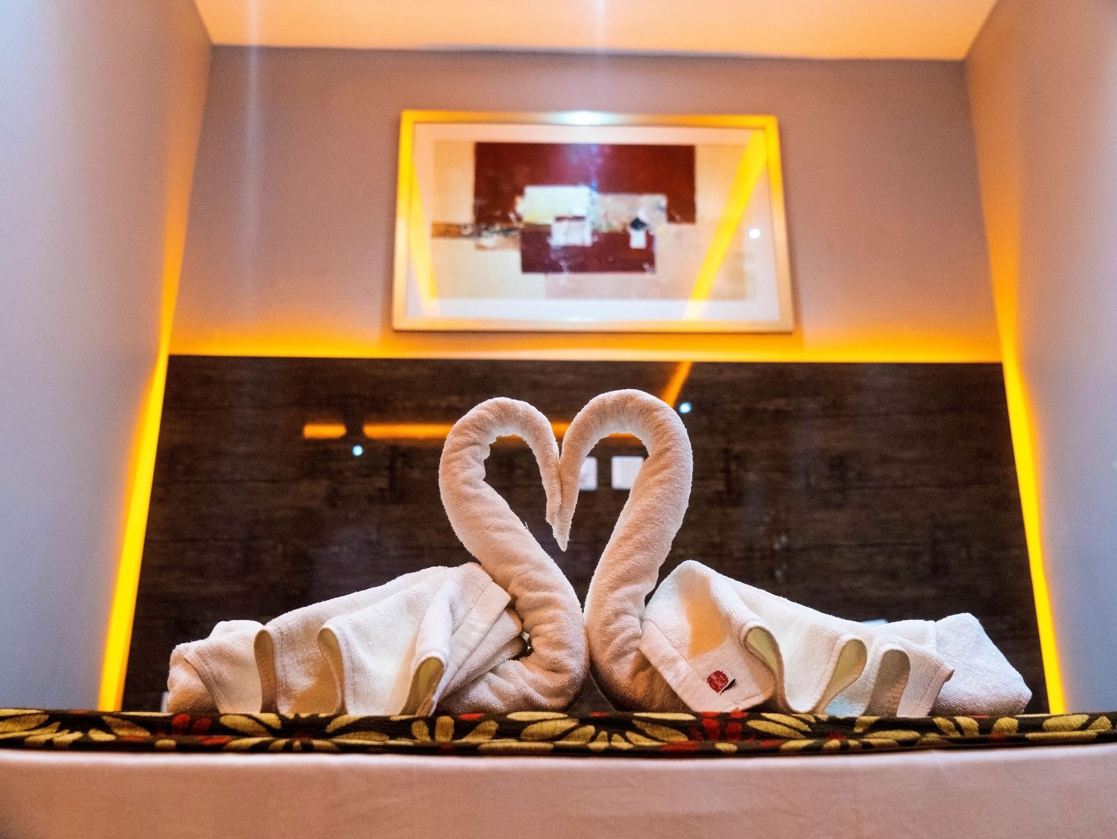 A room with a bed, a framed picture on the wall, and a towel arranged in a heart shape on the bed.