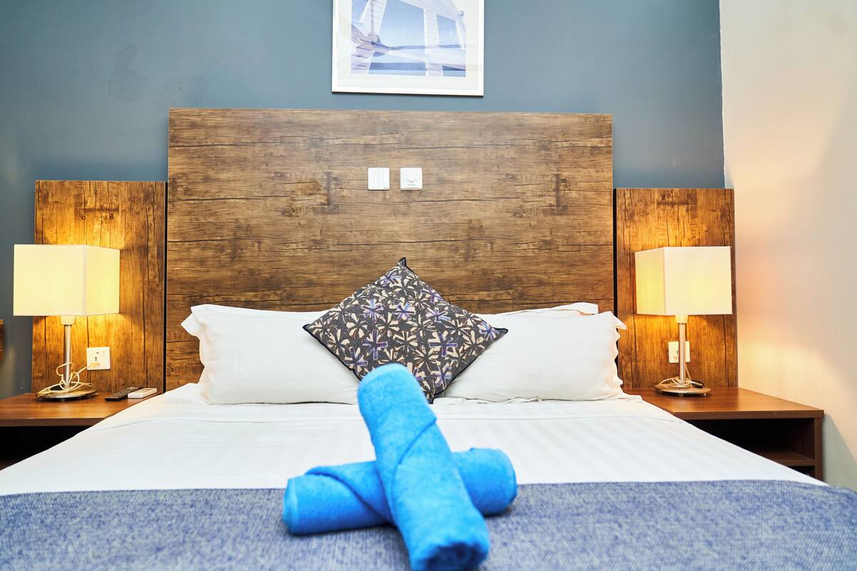 A hotel room with a wooden headboard, two bedside lamps, a bed with white linens, a patterned pillow, and rolled towels on the bed.