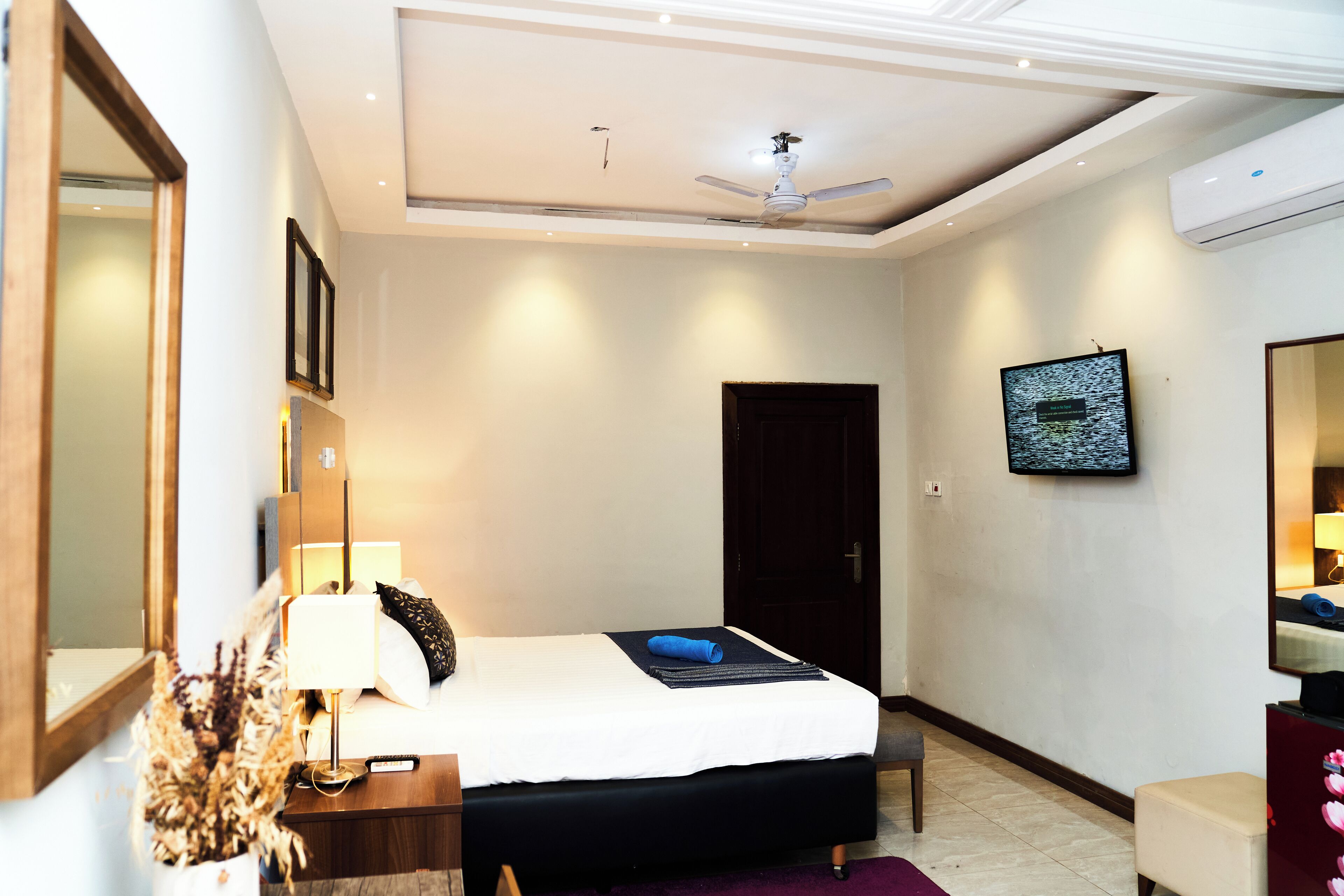 A hotel room with a bed, bedside table, lamp, mirror, TV, and an air conditioning unit.