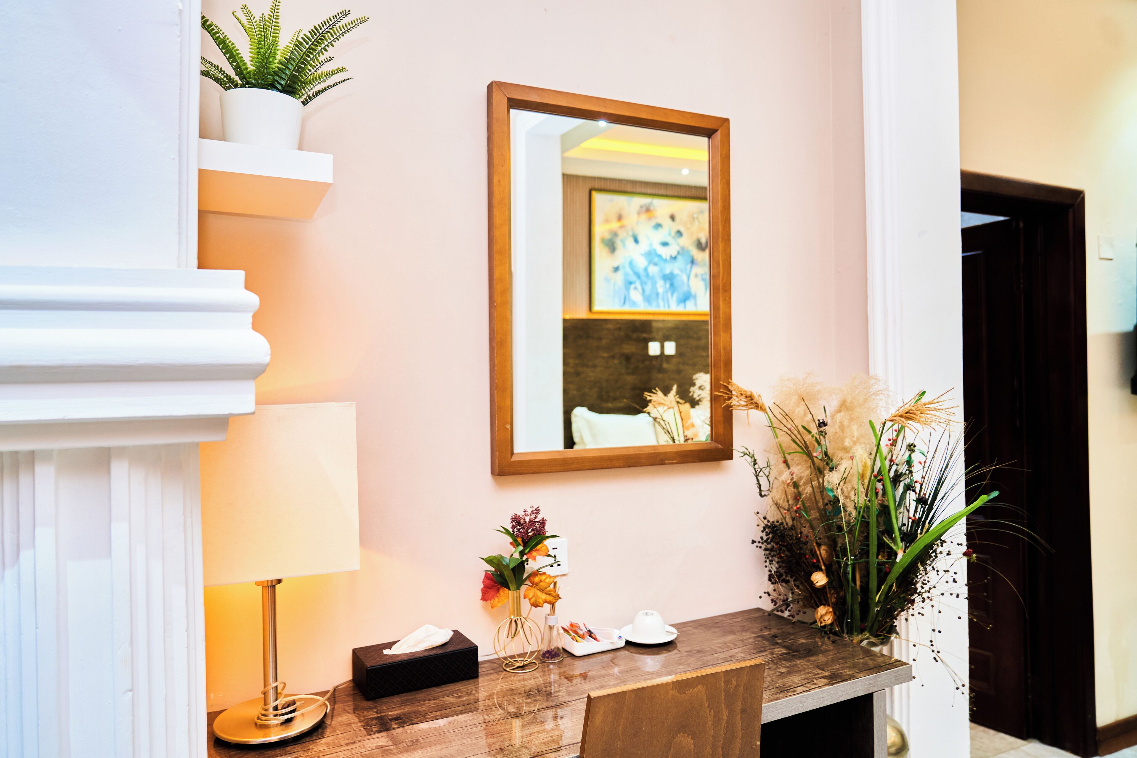 A reception desk with a mirror, a vase with flowers, a lamp, and a small plant on a shelf.