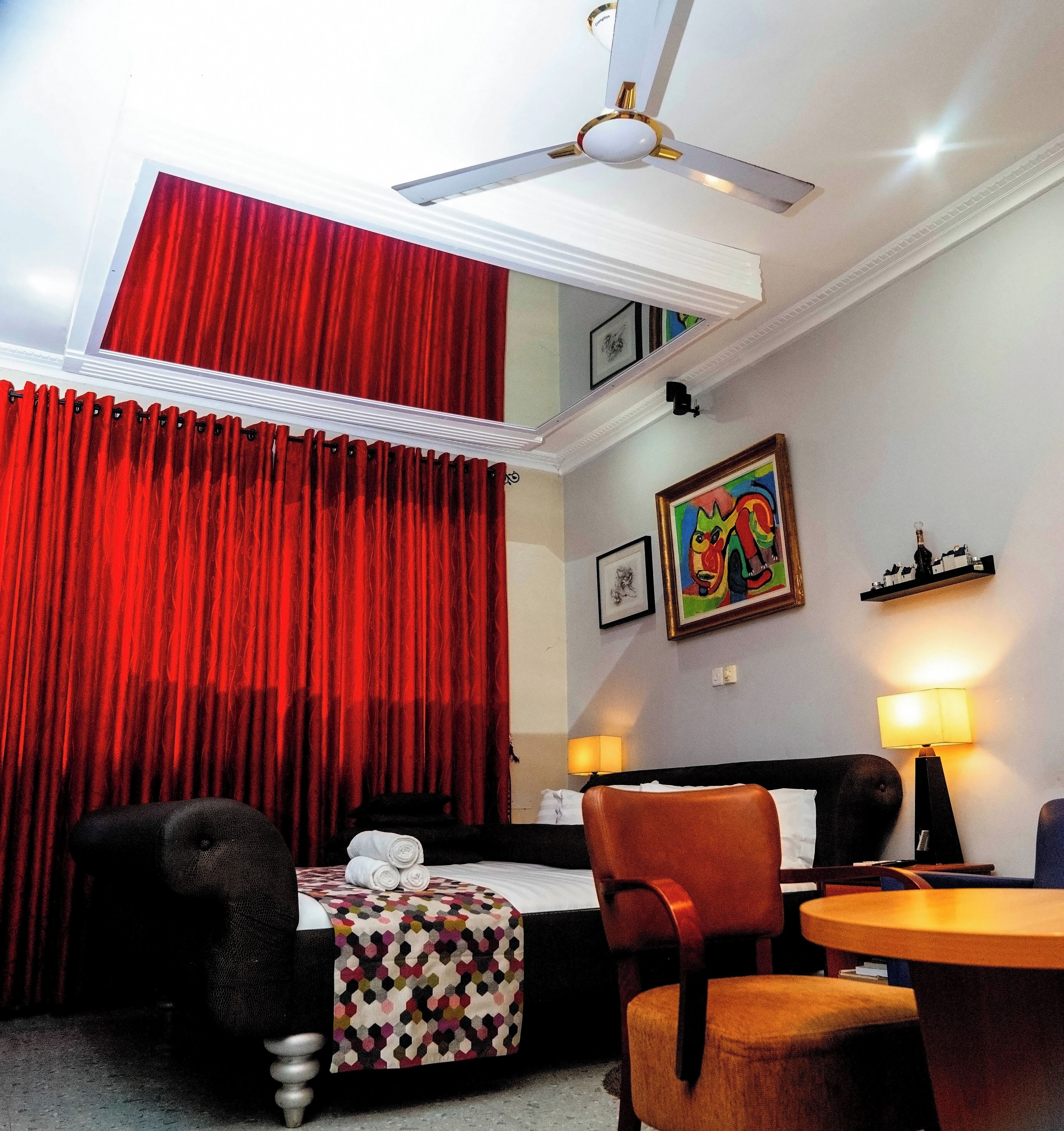 A hotel room with a bed, a desk, a chair, a red curtain, a ceiling fan, and a painting on the wall.