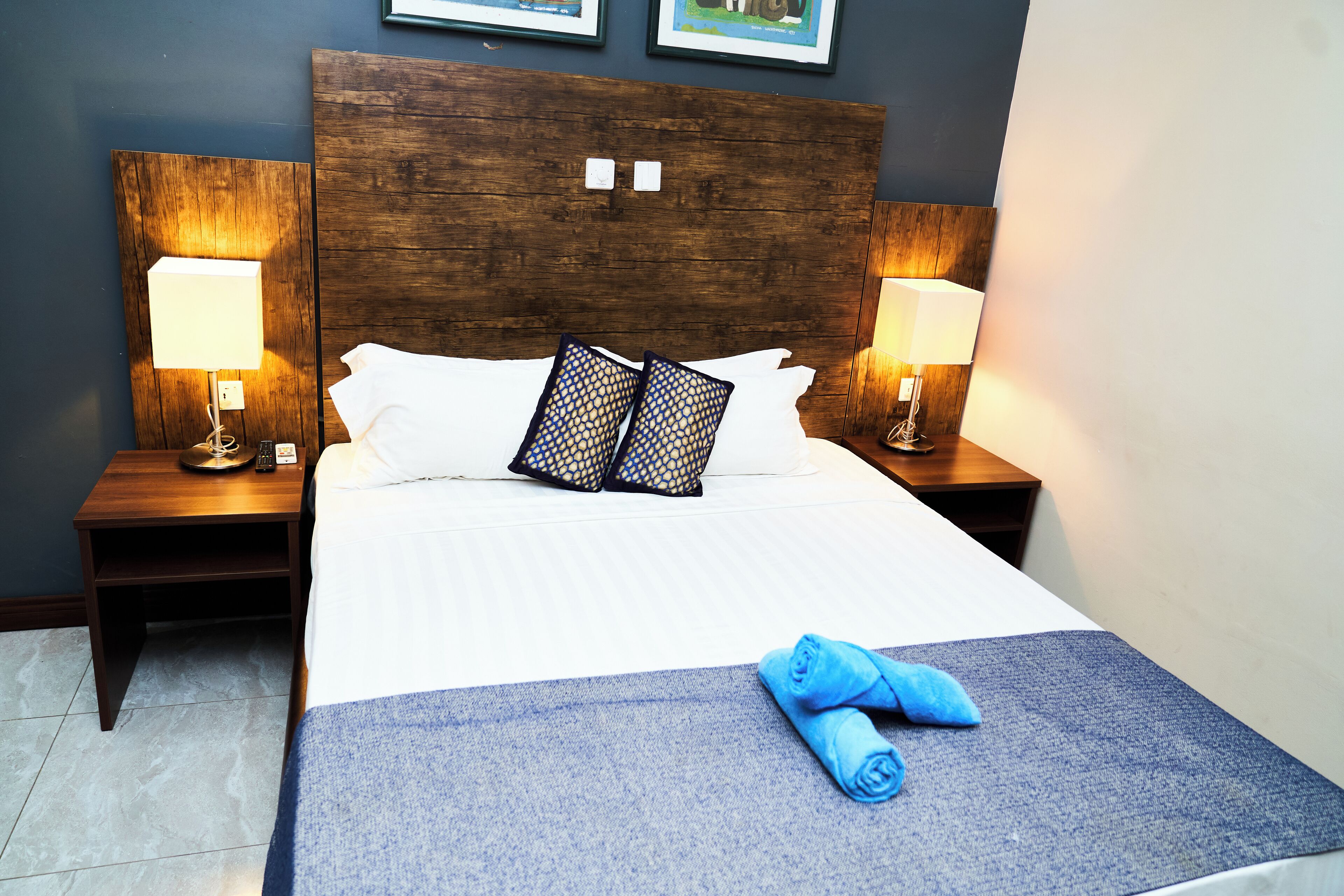 Une chambre d’hôtel avec une tête de lit en bois, deux lampes de chevet, un lit avec du linge de lit blanc, deux coussins décoratifs et une couverture bleue avec des serviettes roulées posées sur le lit.
