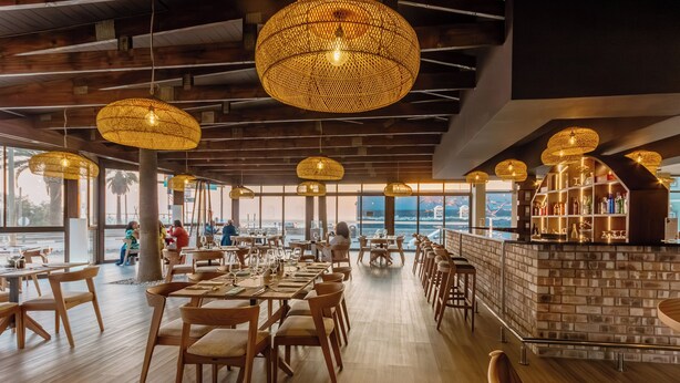 Un restaurant espaiós amb taules i cadires de fusta, llums penjants de fibra natural i vista a l'oceà.