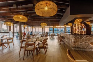 Un restaurante espacioso con mesas y sillas de madera, grandes lámparas colgantes tejidas y vistas al océano.