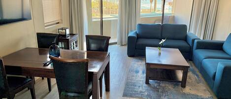 Una sala de estar con sofá, una mesa de centro, una mesa de comedor con sillas y vistas a la playa a través de amplias ventanas.