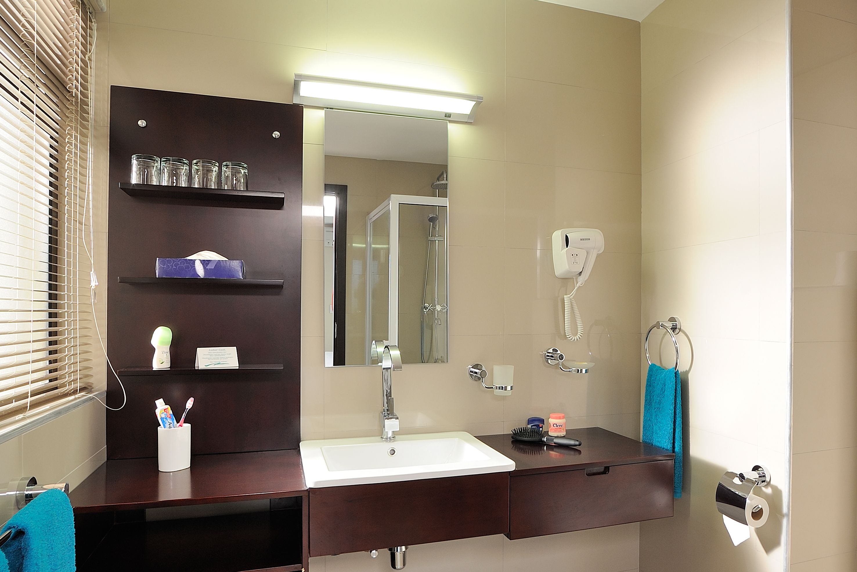 A bathroom with a white sink, dark wooden vanity, a mirror, a towel rack, a hairdryer, and a shelf with toiletries.