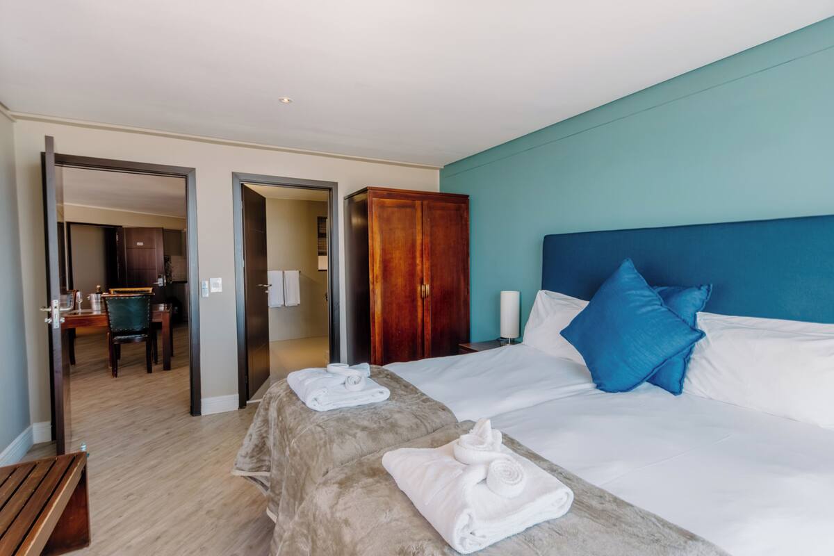 A hotel room with a large bed, a wooden wardrobe, a desk with a chair, and a bathroom visible through an open door.