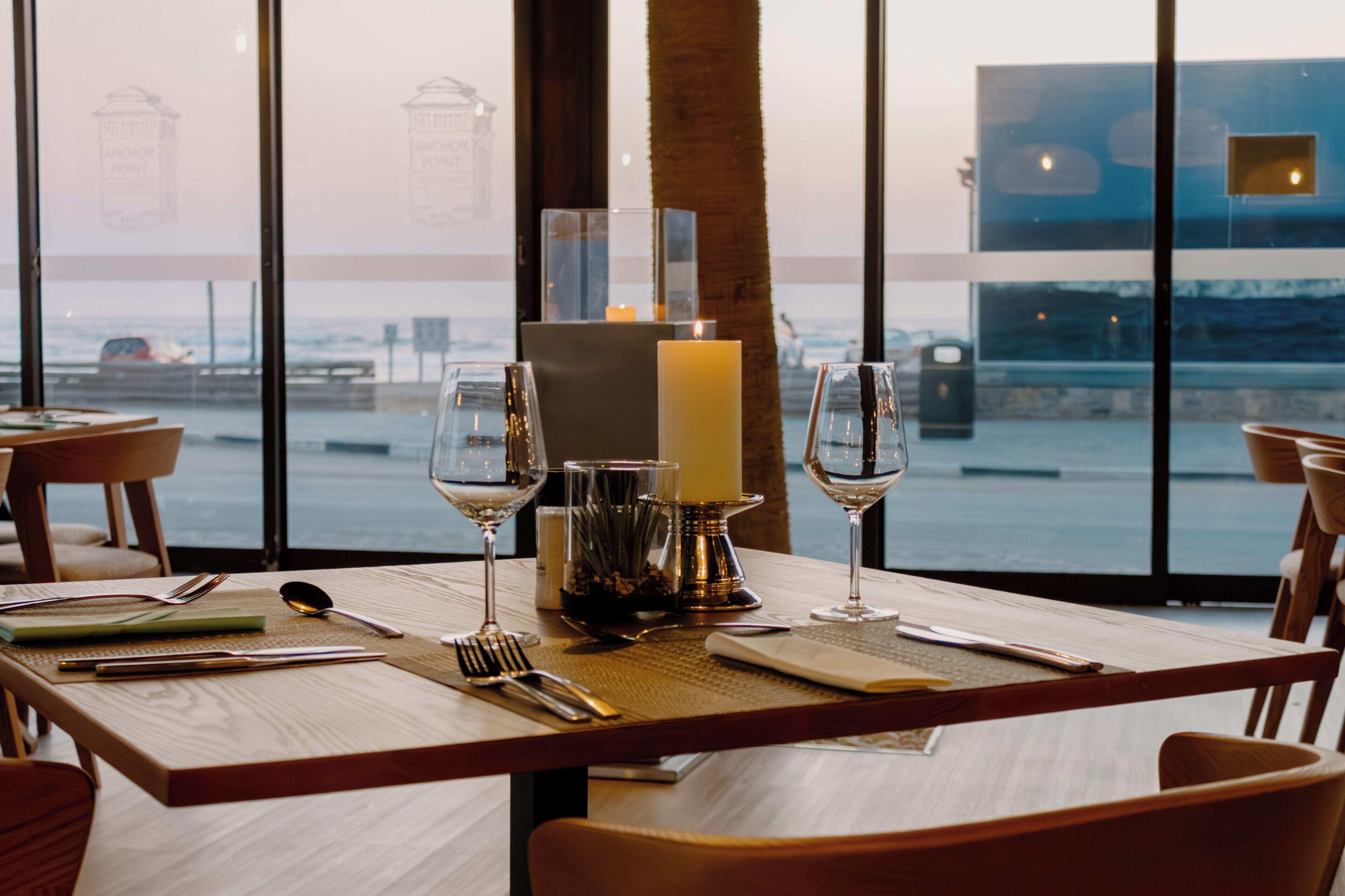 Une table à manger dressée avec une bougie, des verres à vin et des couverts, offrant une vue sur la mer et un bâtiment.