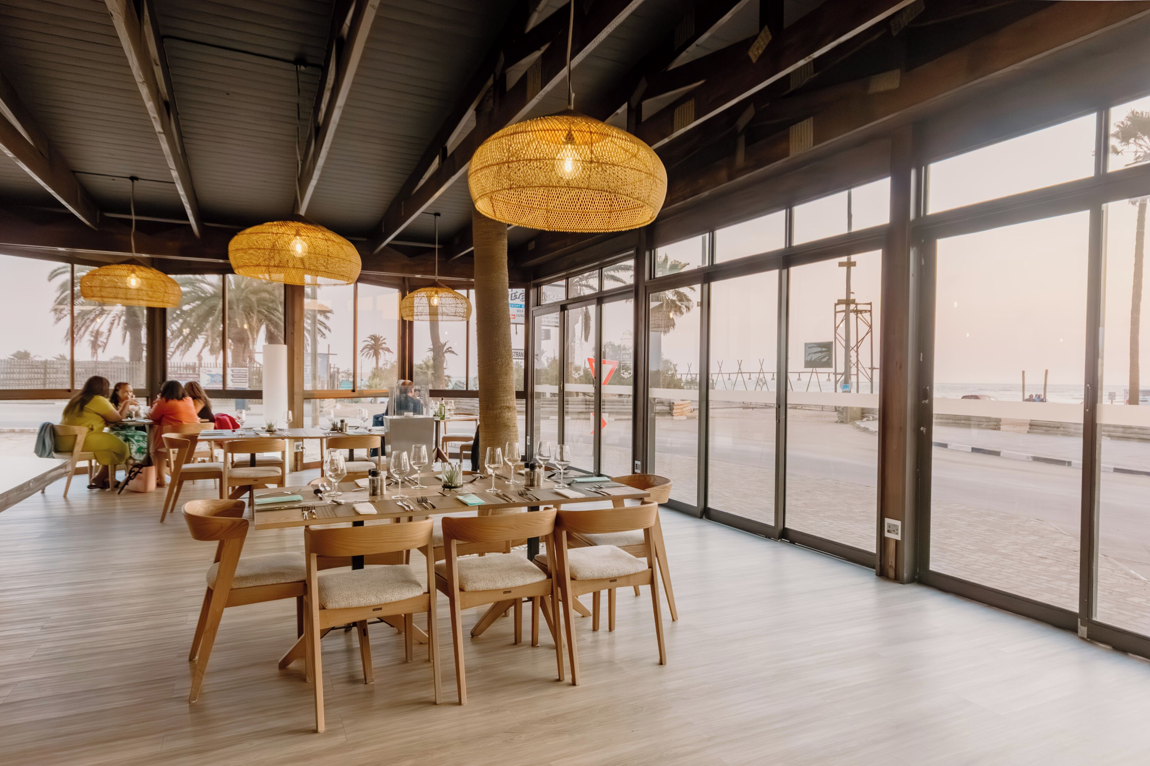 A restaurant with a large dining area, wooden tables and chairs, and large windows offering a view of the beach.