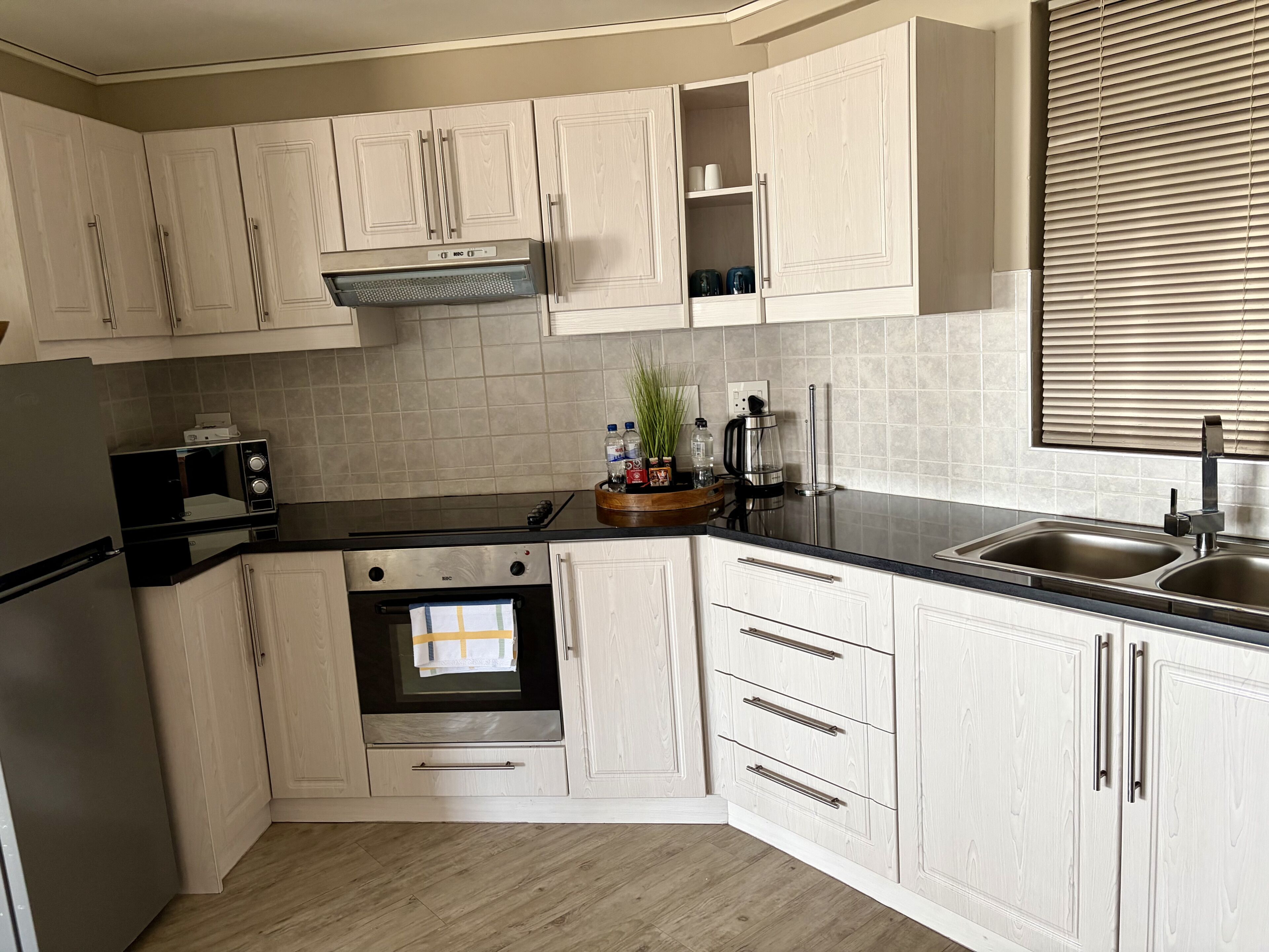 Een keuken met witte kastjes, een roestvrijstalen koelkast, een magnetron, een oven, een spoelbak en een raam met jaloezieën.
