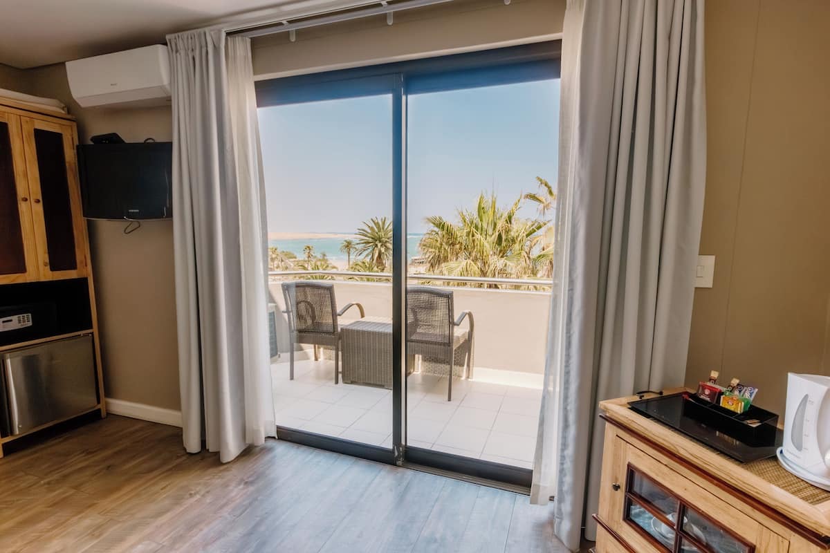A room with a sliding glass door leading to a balcony with a view of the beach and palm trees.
