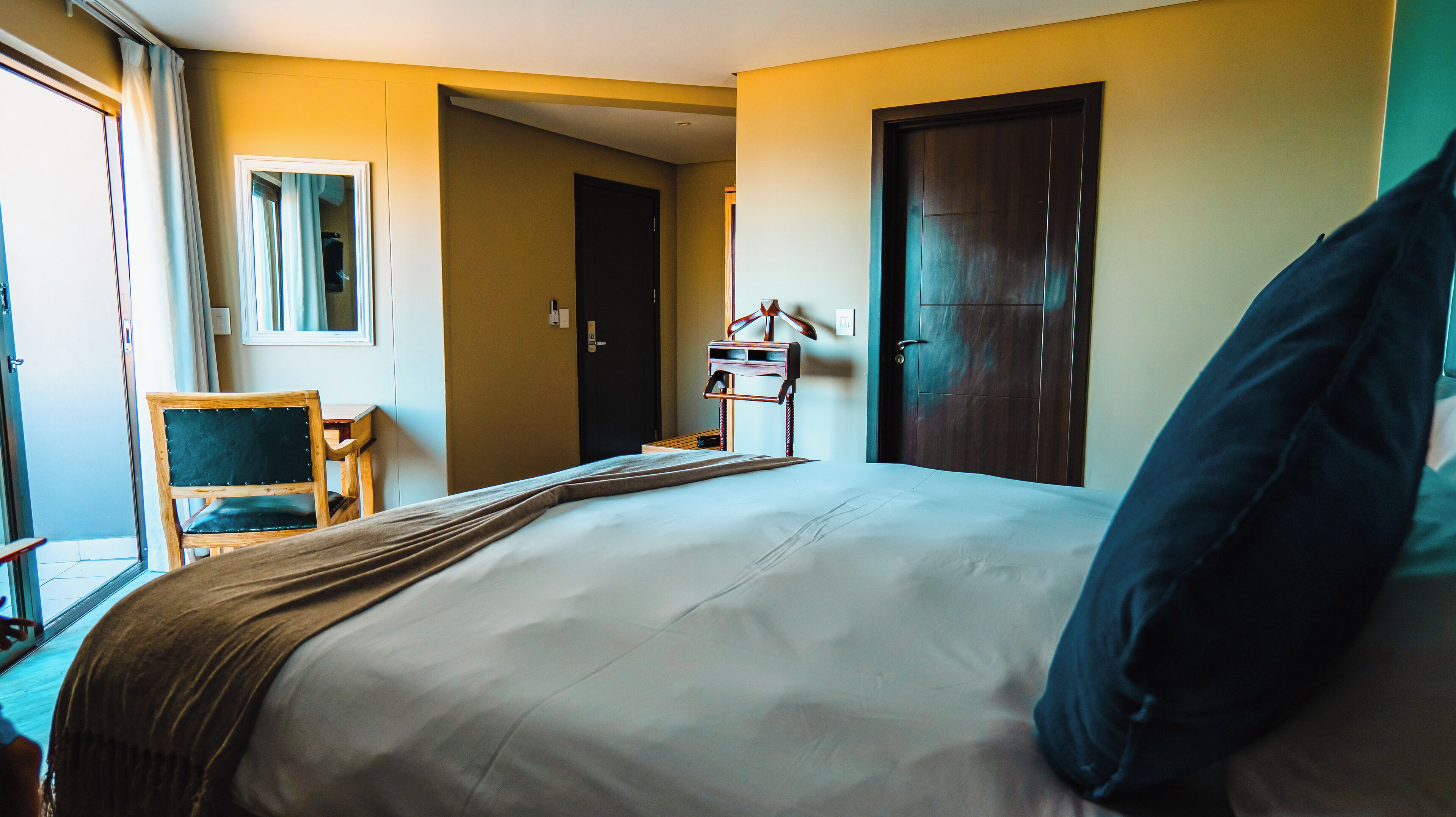 Habitación de hotel con una cama grande, un escritorio y una silla. Se observa una puerta y una ventana con cortinas.