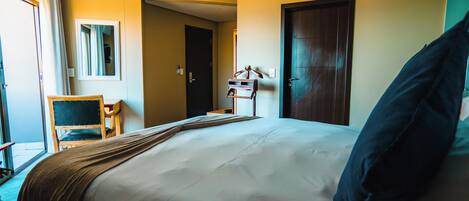 Habitación de hotel con una cama grande, un escritorio y una silla. Se observa una puerta y una ventana con cortinas.