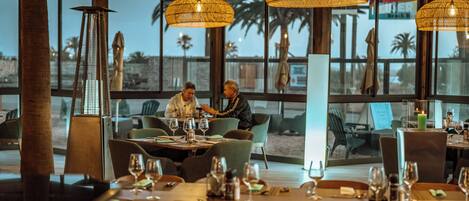 A restaurant with tables set for dining, large windows, and hanging wicker lamps.