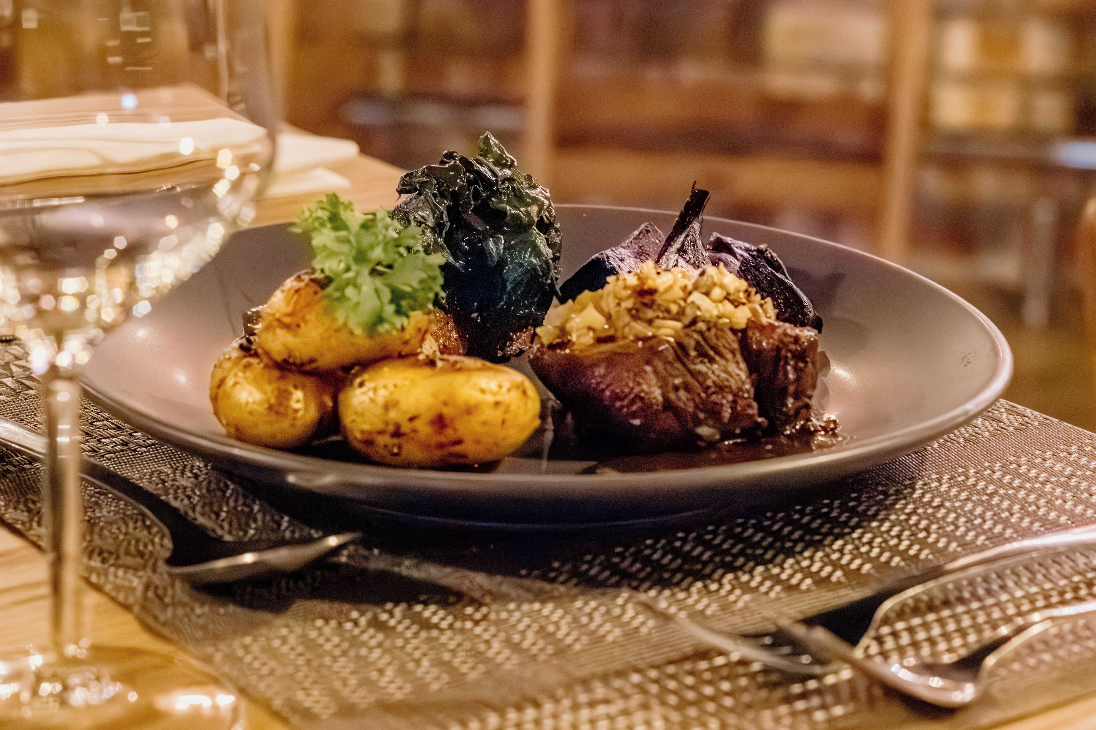 Un plato de comida con patatas, verduras y carne, acompañado de una copa de vino sobre una mesa con posavasos.