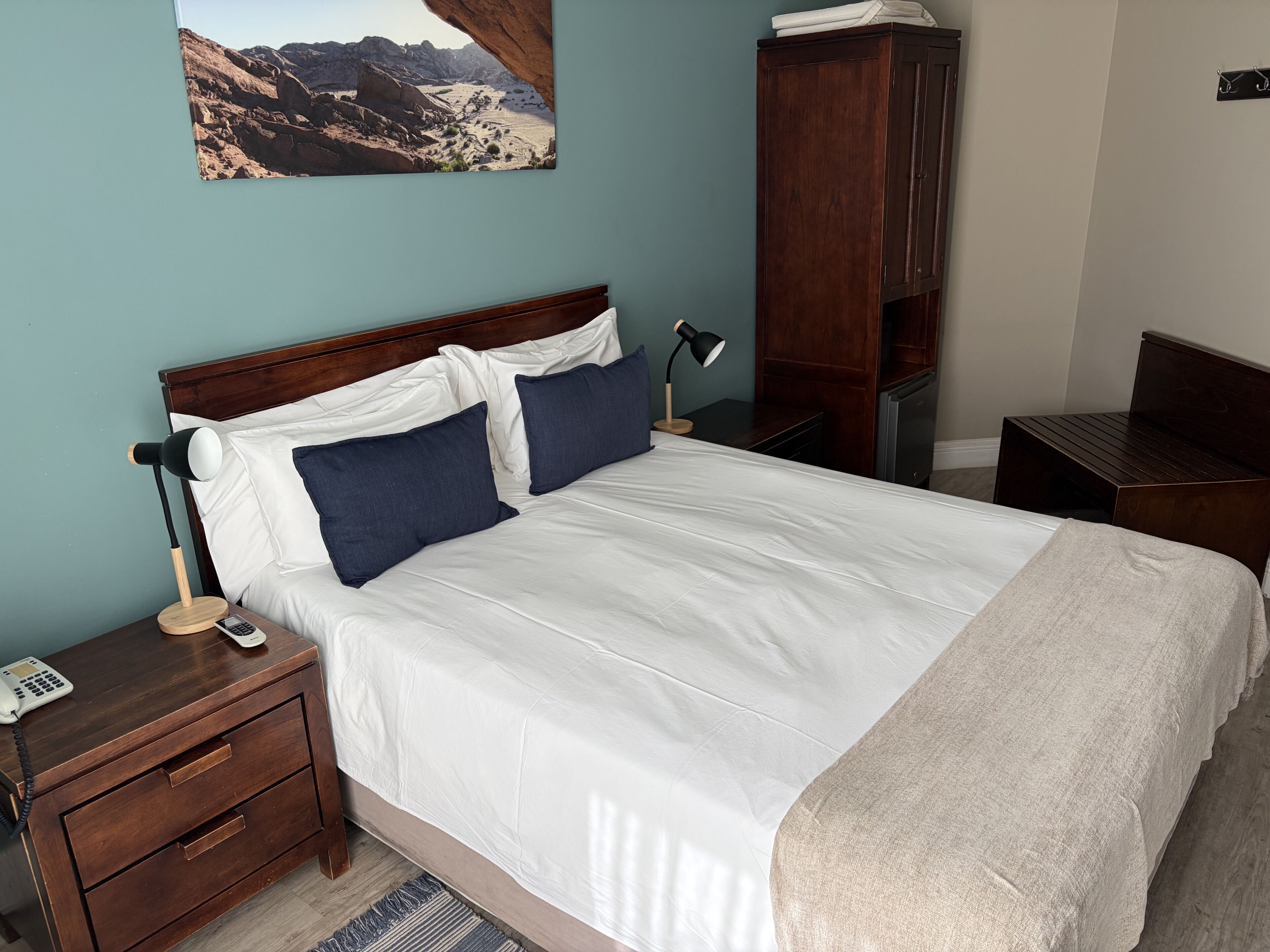 Un dormitorio con una cama, una mesita de noche, una lámpara, un teléfono, un armario de madera y un cuadro de un paisaje.