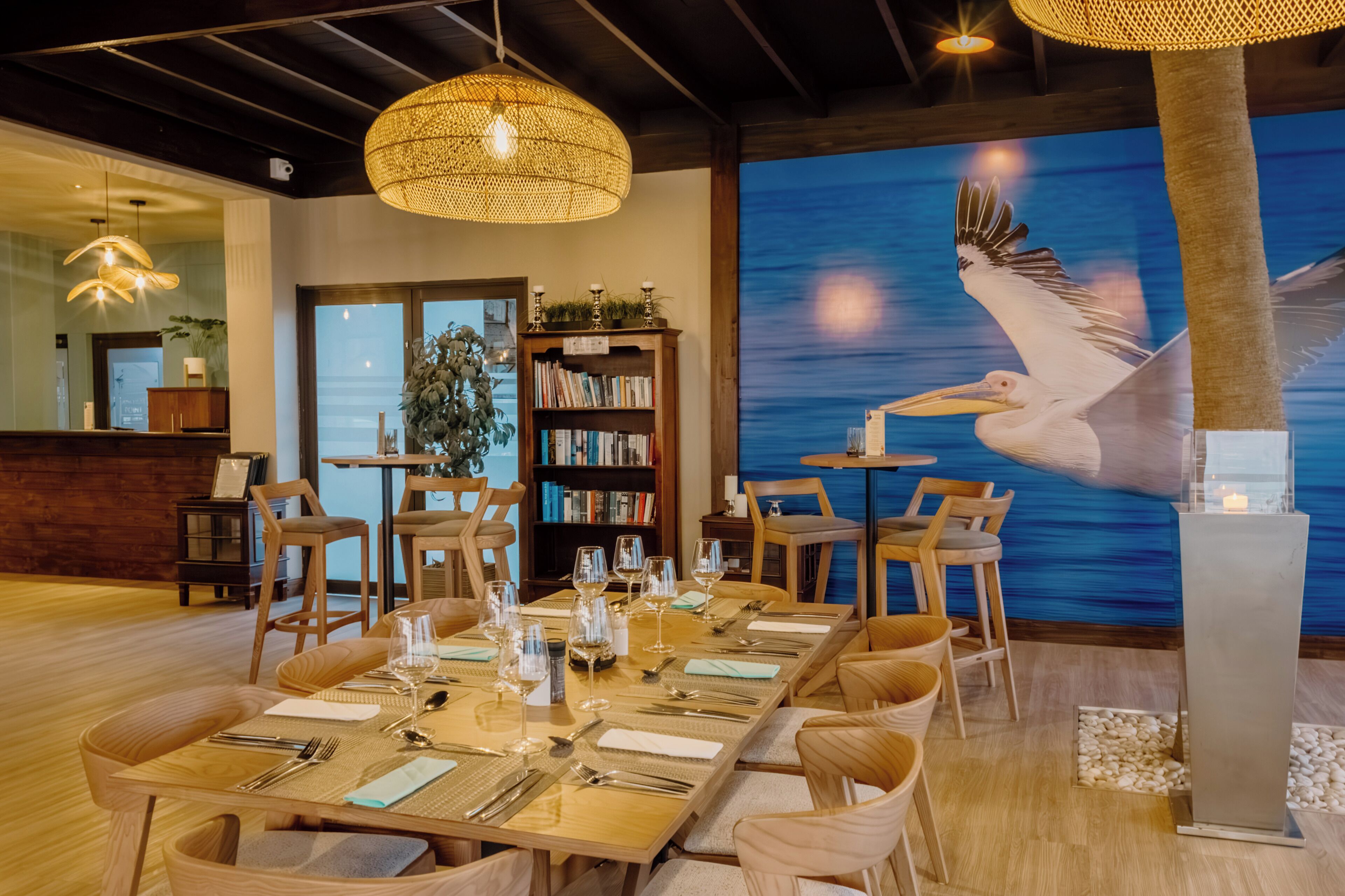 Un coin repas avec une longue table dressée pour six personnes, entourée de chaises en bois. On y trouve une bibliothèque remplie de livres et une plante décorative. Un grand mur peint représente un oiseau.