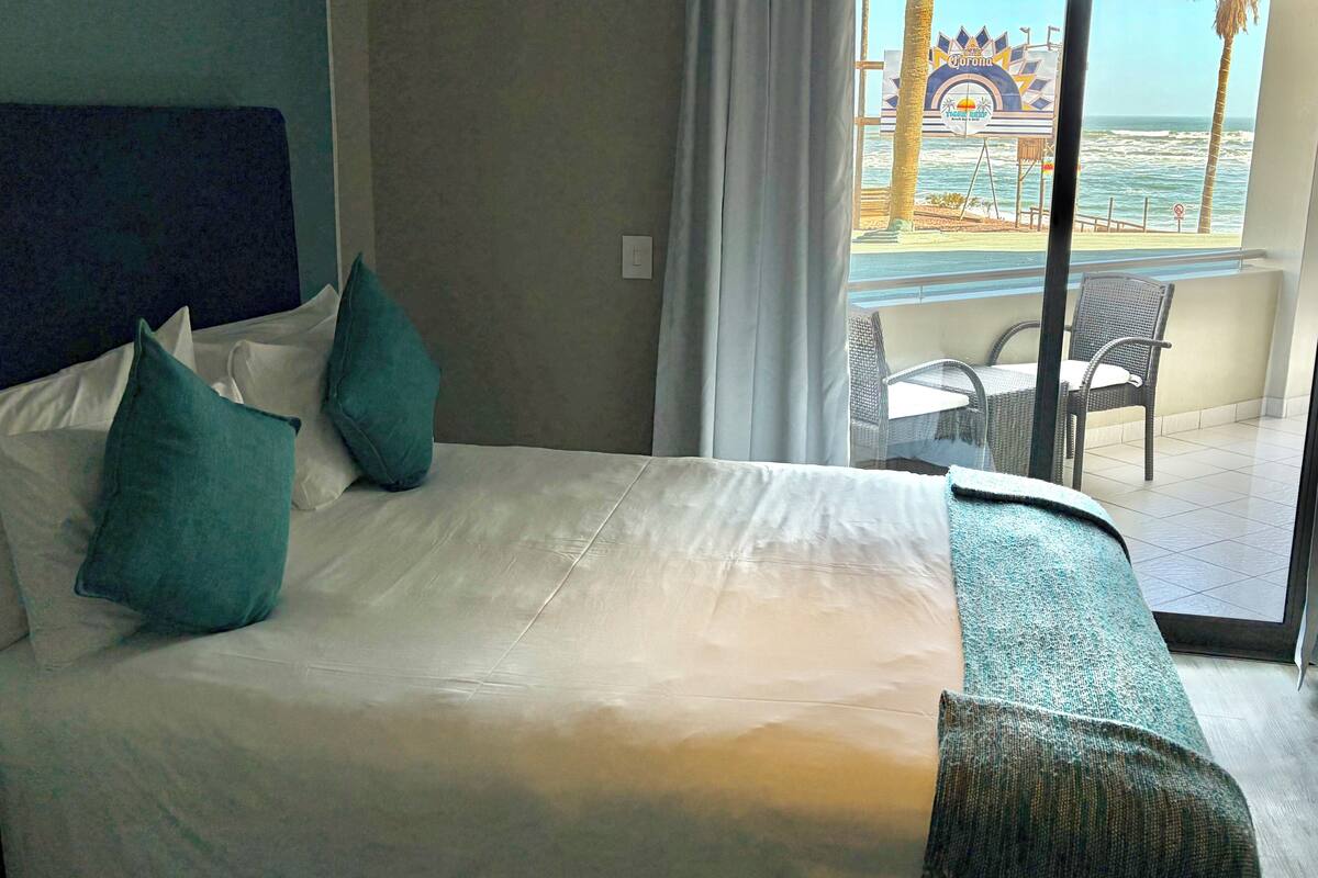 A hotel room with a bed, pillows, a window with a view of the beach and a sign, and a balcony with chairs and a table.
