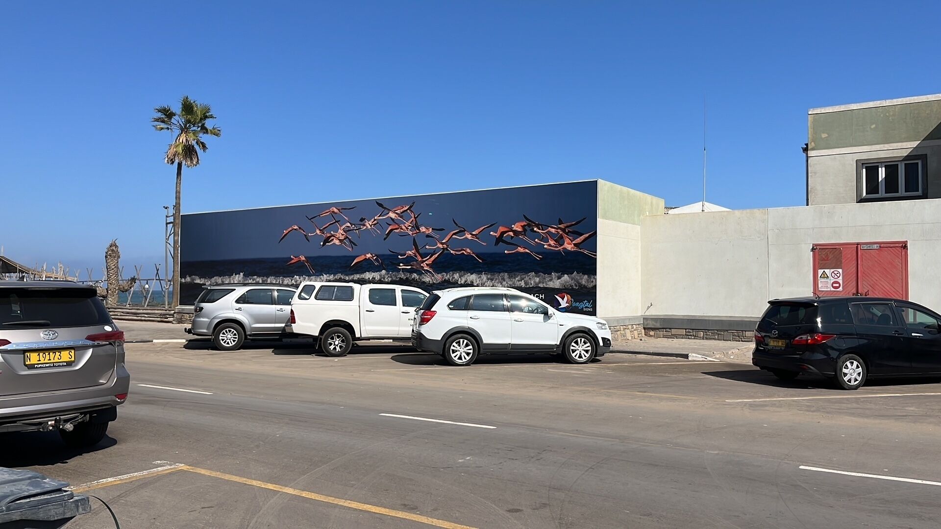 A parking lot with several cars, a mural of flamingos on a building, and a palm tree in the background.