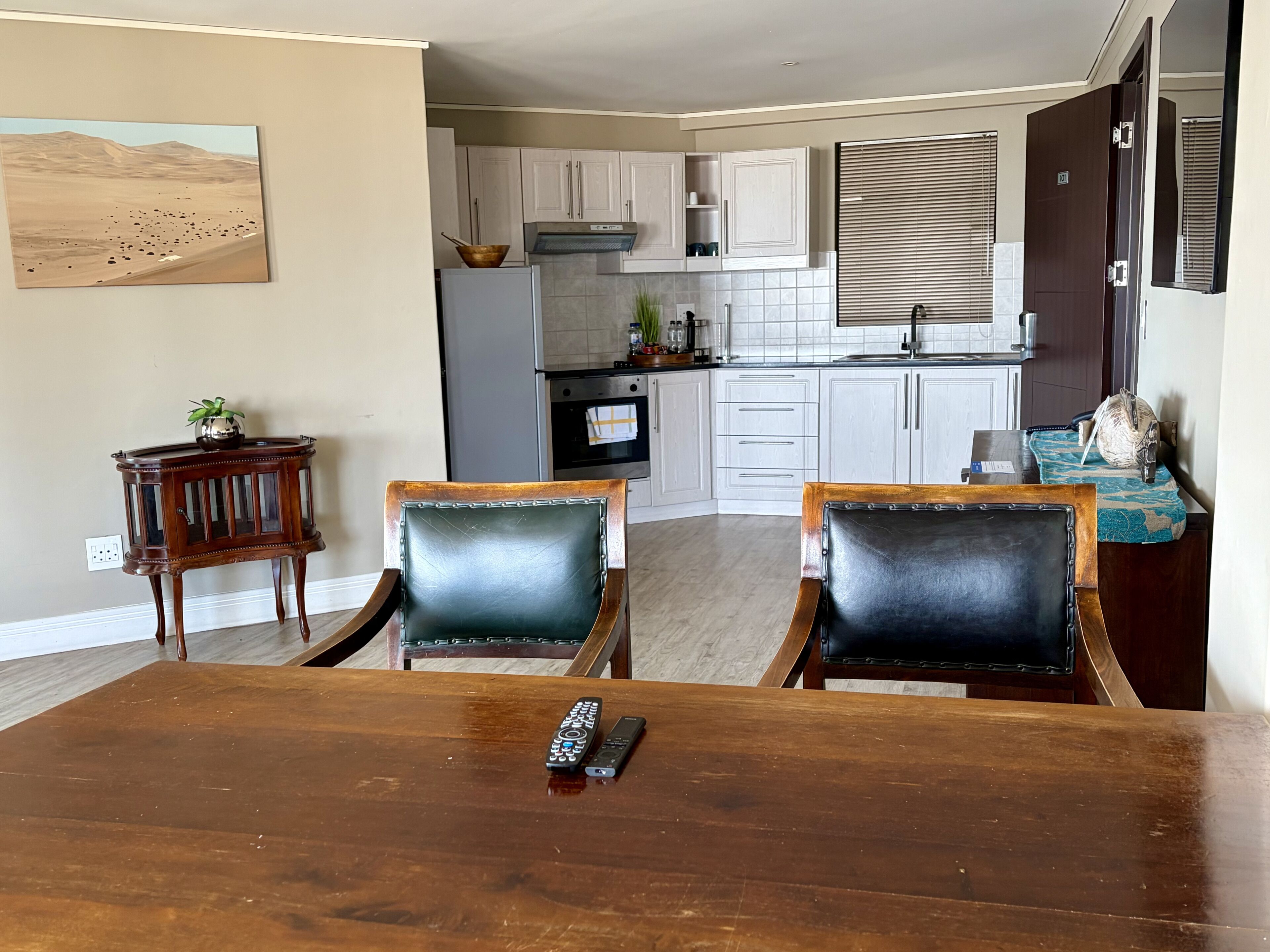 A dining area with a wooden table and chairs, a remote control on the table, and a kitchen in the background.