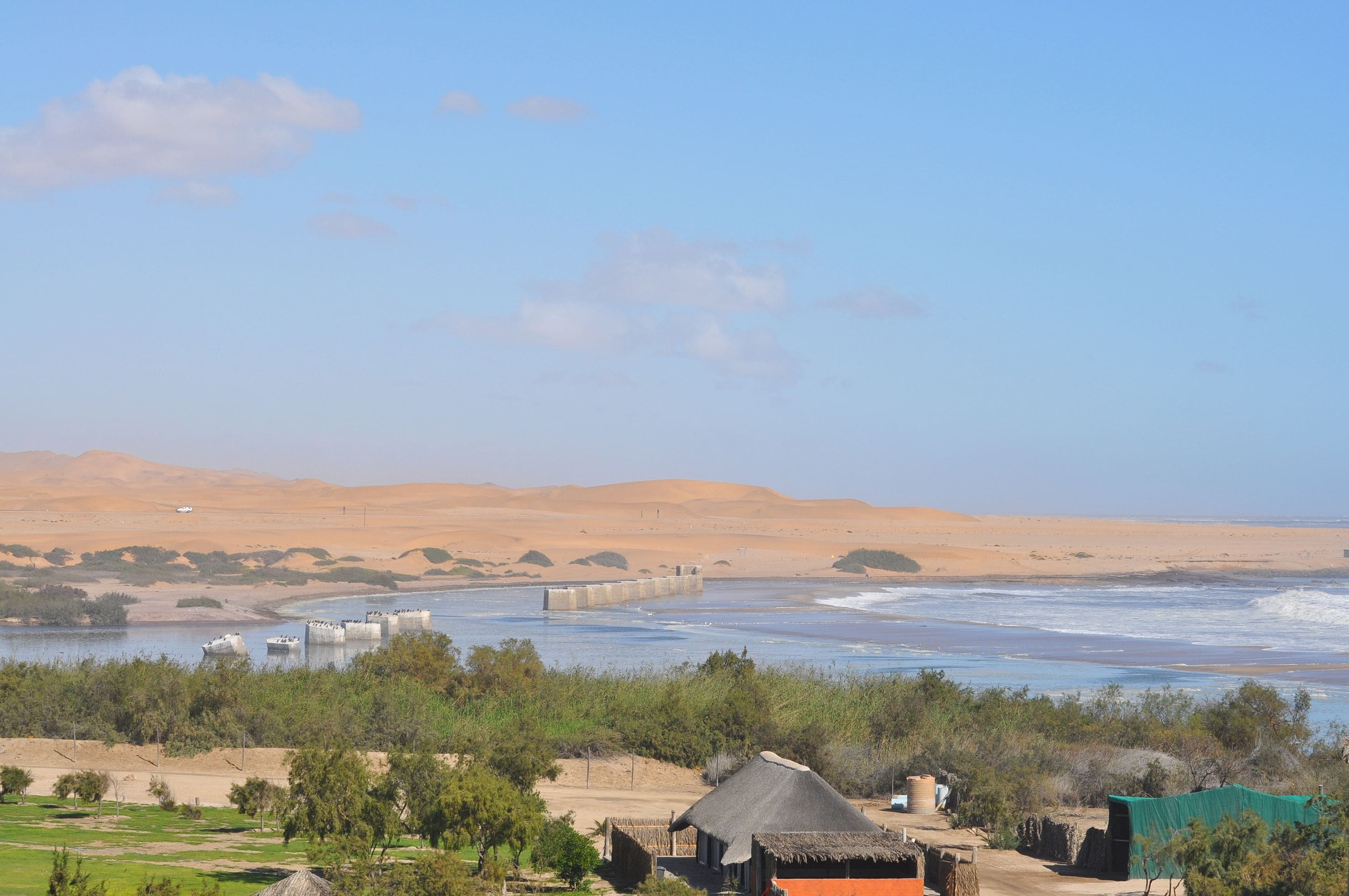 A coastal landscape with sand dunes, a body of water, and a small settlement with thatched-roof structures.
