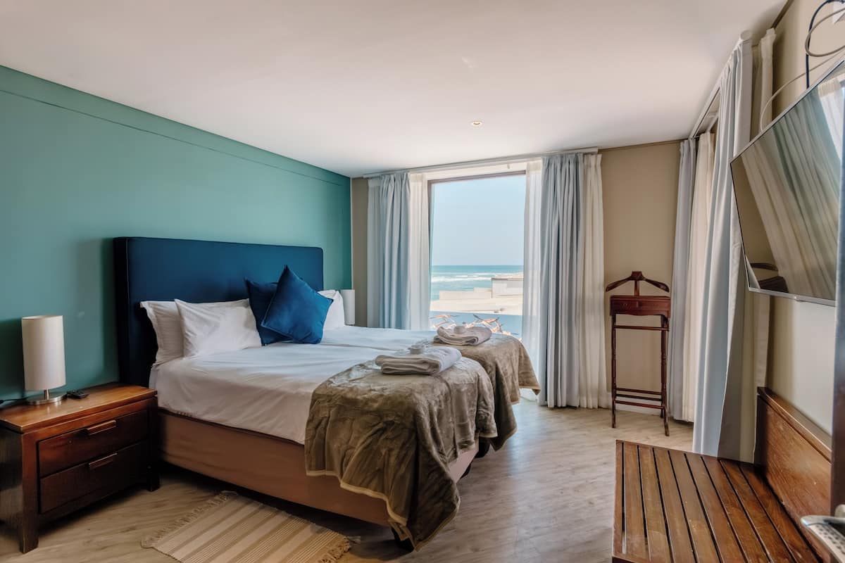 A hotel room with a large bed, a wooden bench, a nightstand, and a view of the beach.