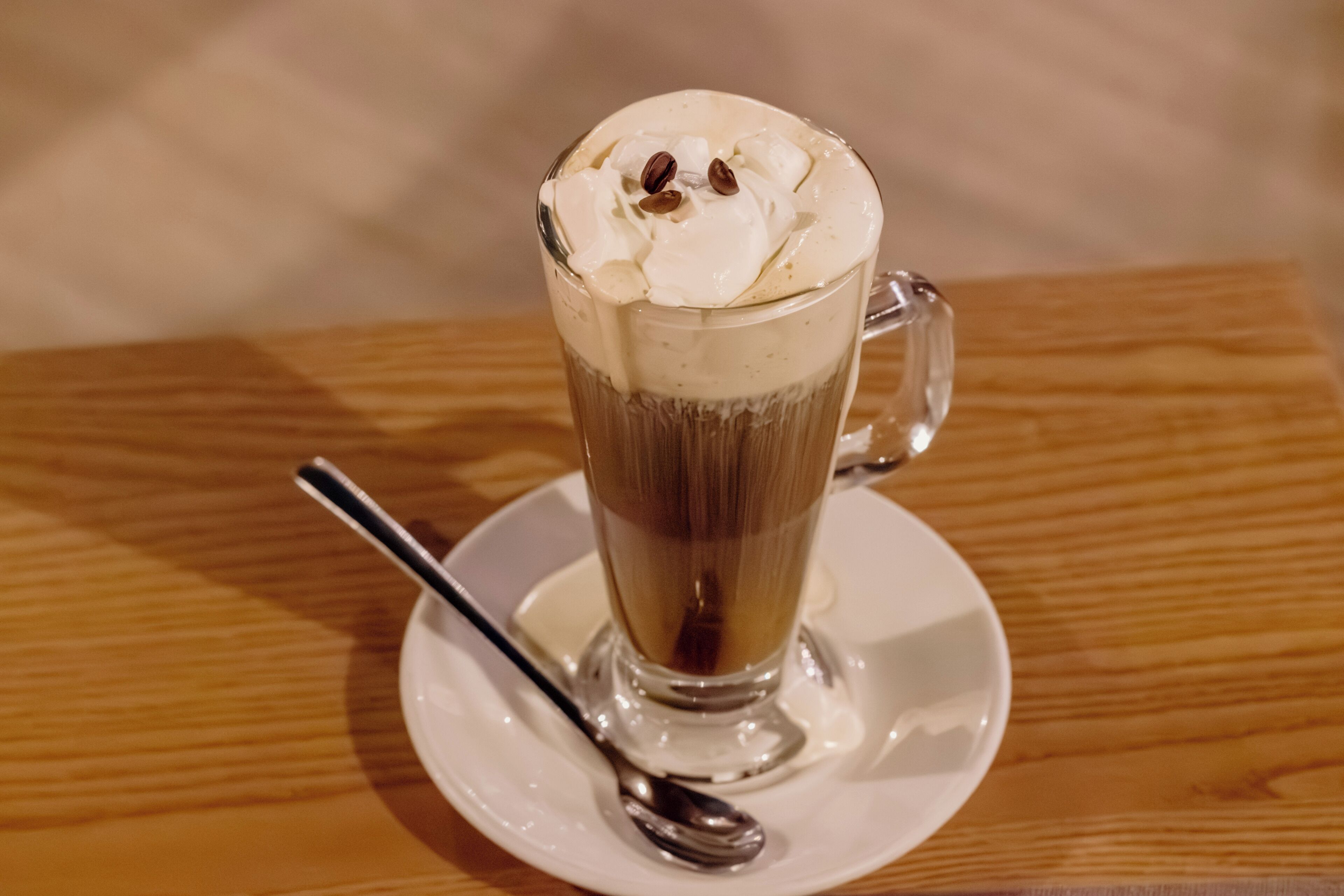 A tall glass mug with whipped cream and coffee beans on top, placed on a white saucer with a spoon.