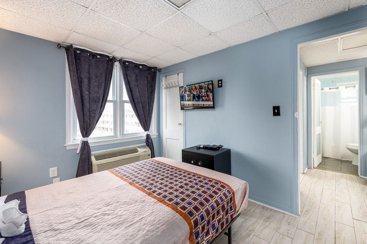 A bedroom with a bed, a window with curtains, a wall-mounted TV, a small black cabinet, and a door leading to a bathroom.