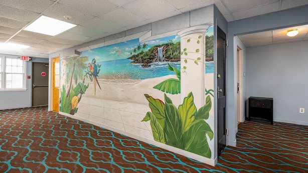 A hotel lobby with a mural of a beach scene, a patterned carpet, and a door with an exit sign. - Hammock Inn & Suites - Jersey Shore (Seaside Heights)