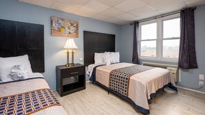 A hotel room with two beds, a nightstand with a lamp, a window with curtains, and a wall art piece. - Hammock Inn & Suites - Jersey Shore (Seaside Heights)
