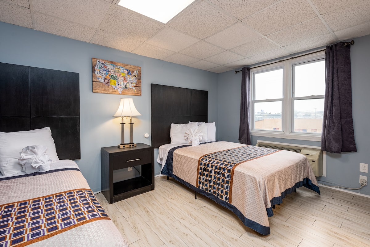 A hotel room with two beds, a nightstand with a lamp, a window with curtains, and a wall art piece.