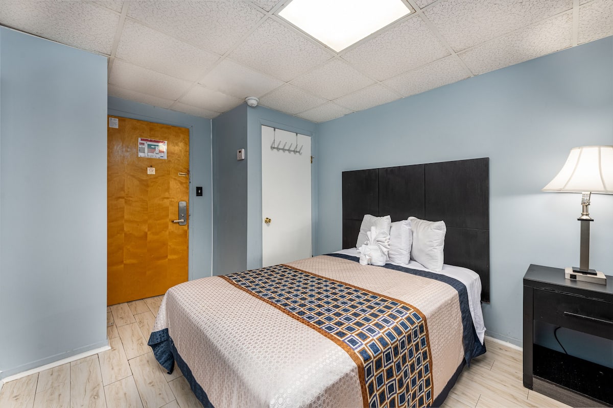 A hotel room with a bed, a nightstand, a lamp, a door, and a ceiling light.