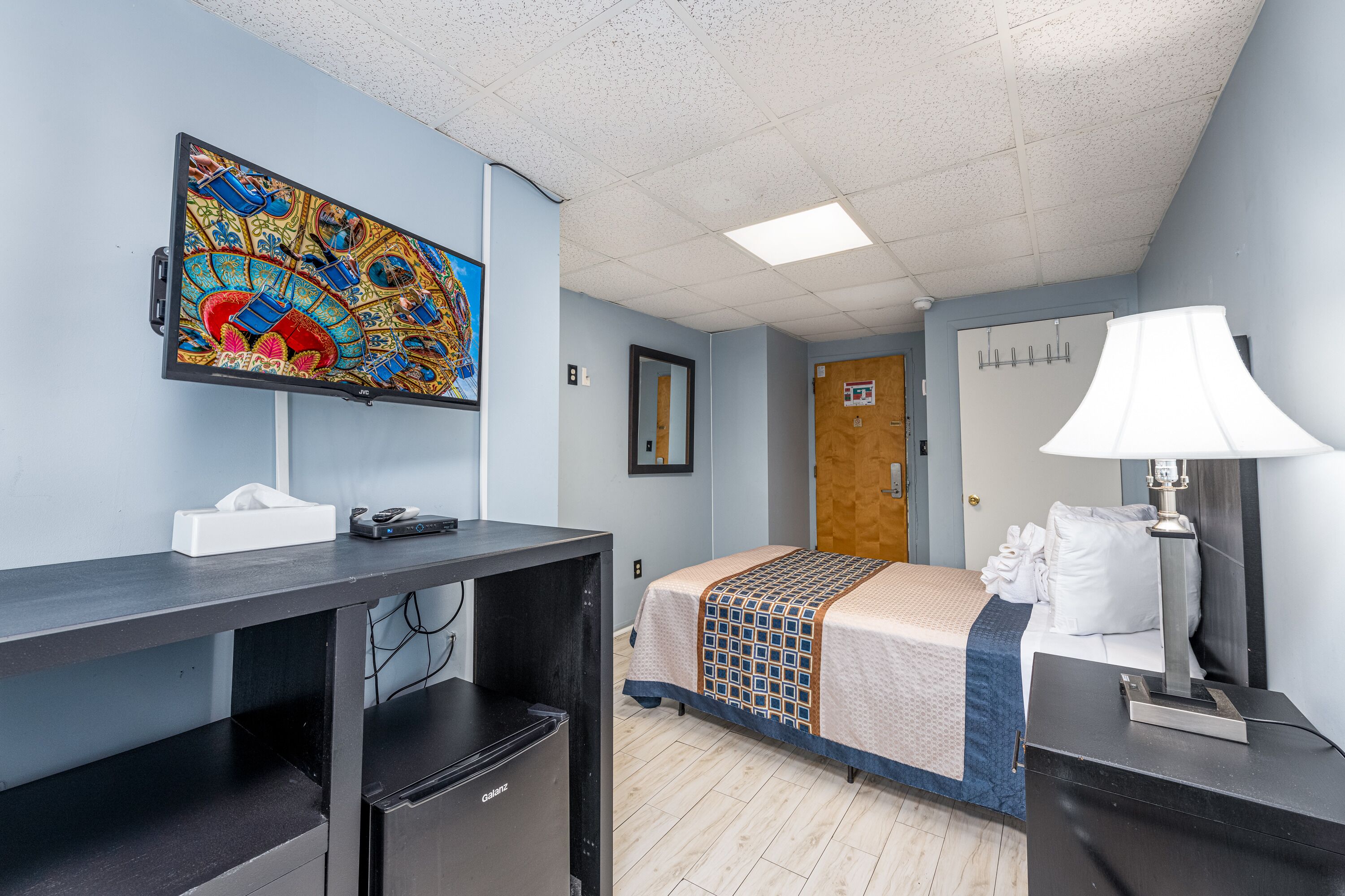 Una habitación de hotel con una cama, un escritorio con una computadora, una lámpara y una obra de arte colorida en la pared.