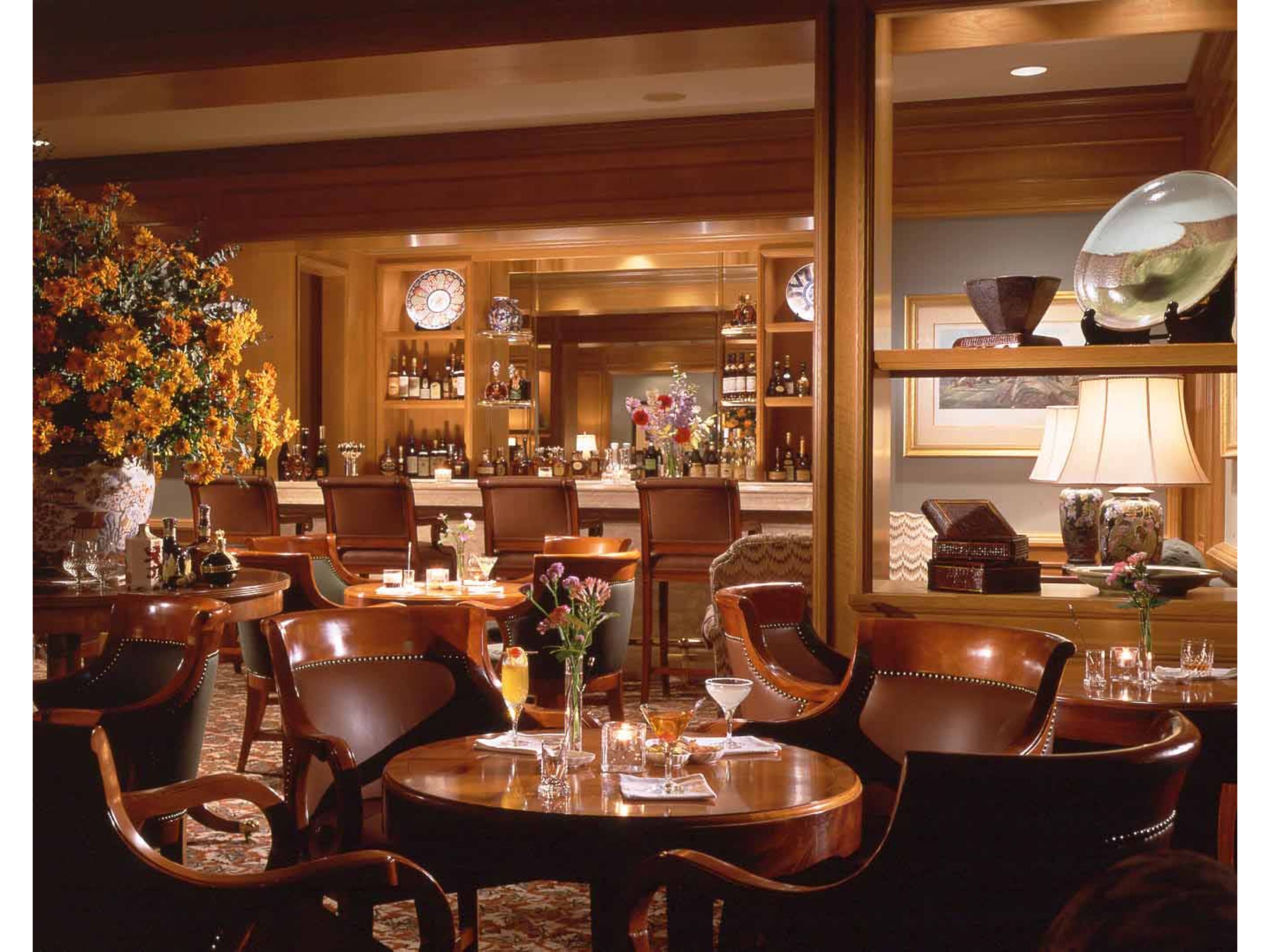 Un elegante bar bien iluminado con mobiliario de madera, un gran arreglo floral y una variedad de botellas y copas en estantes.