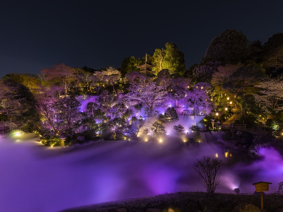 Escena nocturna amb una cascada, arbres il·luminats i arquitectura tradicional.