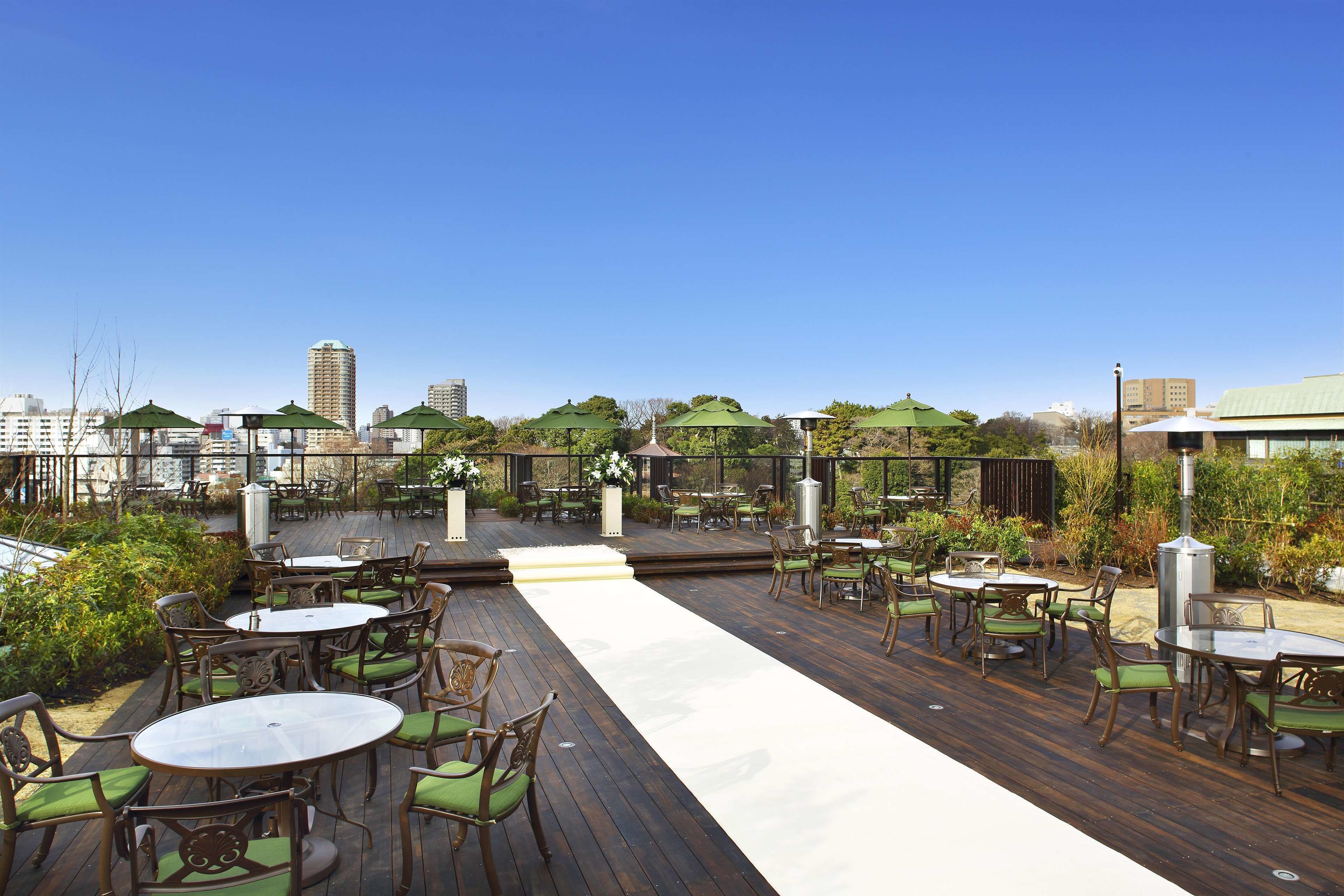 An outdoor patio with wooden decking, green cushioned chairs, and tables, overlooking a cityscape.