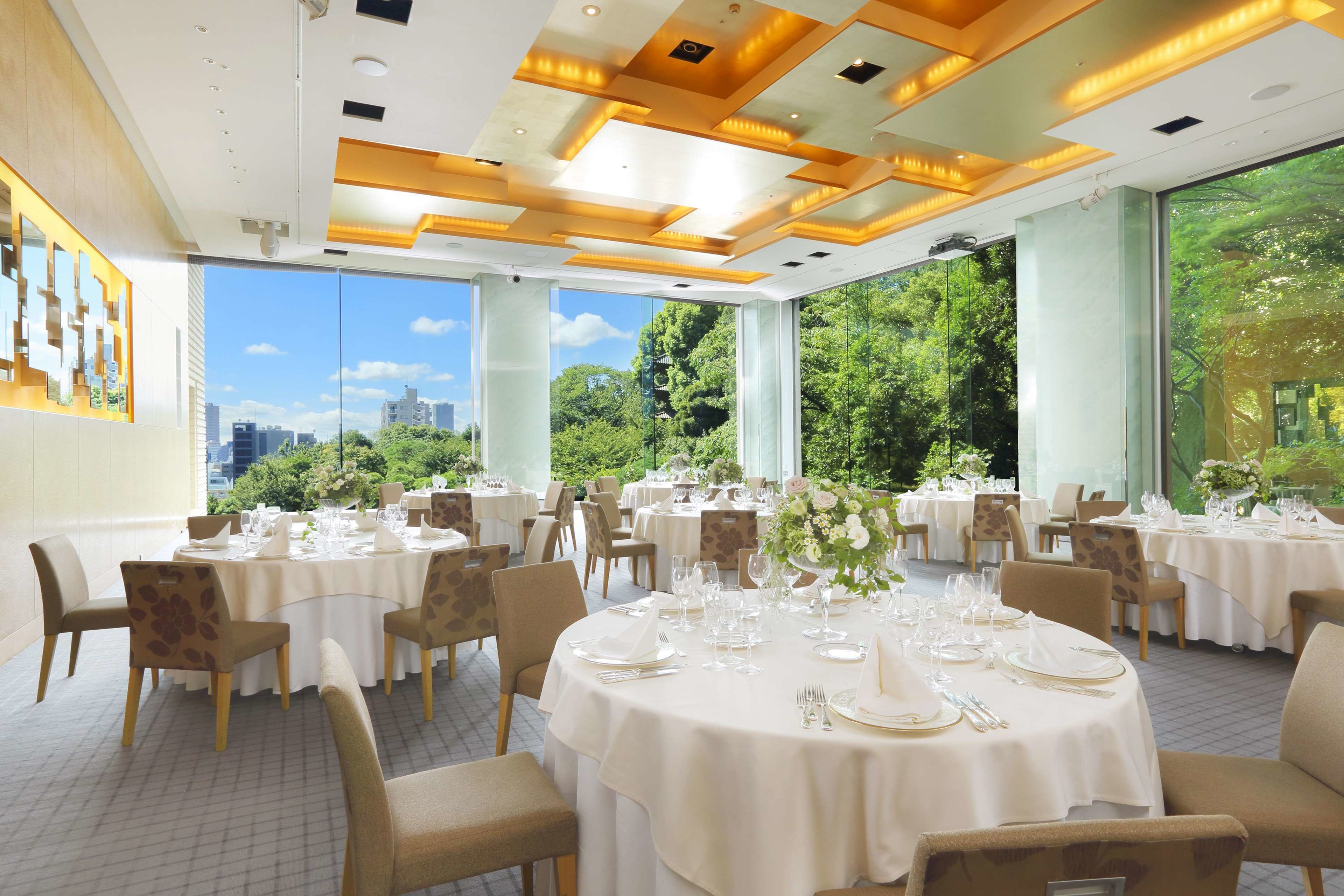A dining area with round tables set for a formal meal, large windows offering a view of greenery and buildings, and a modern ceiling design.