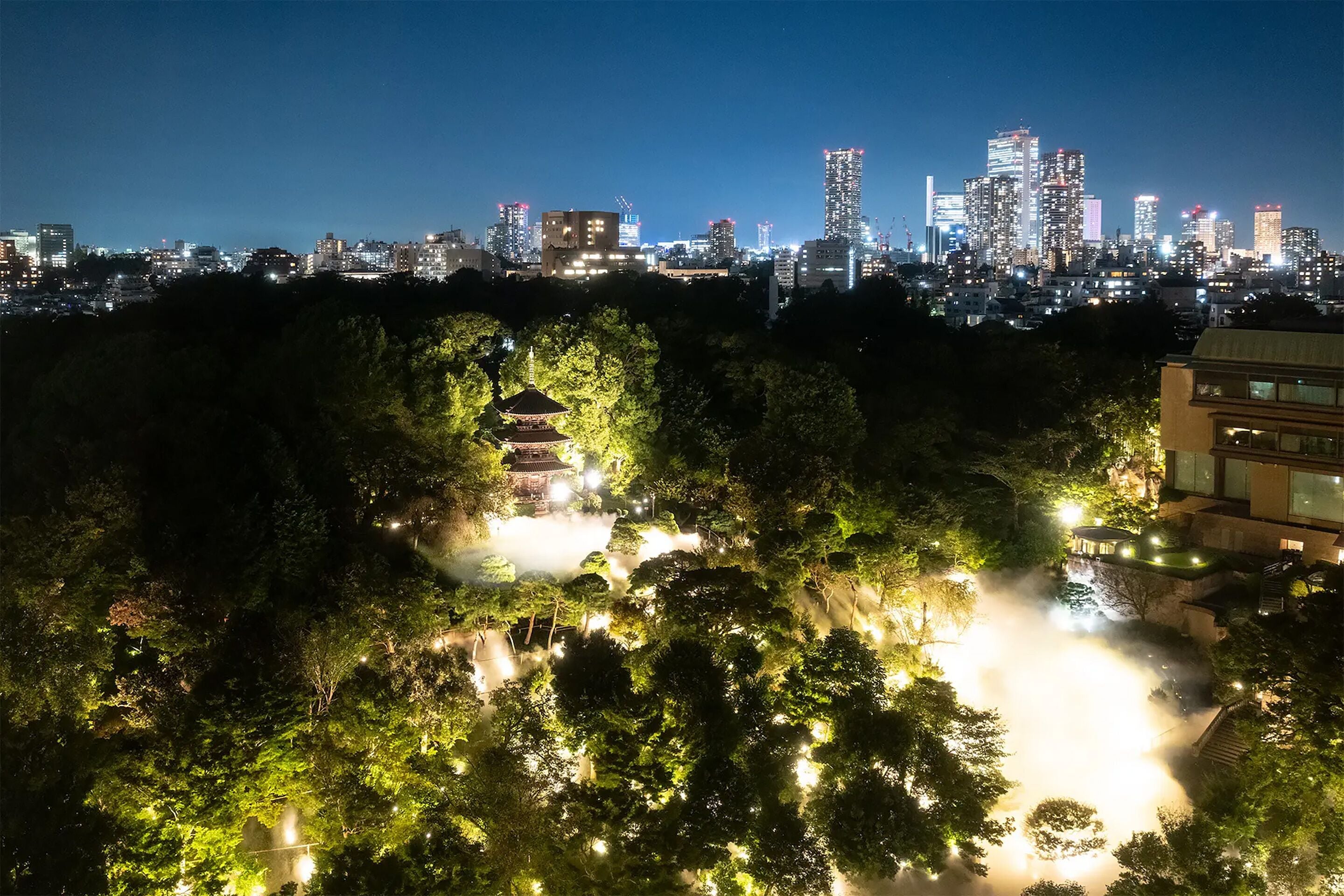 A cityscape at night with a park area featuring a traditional pagoda and illuminated pathways.