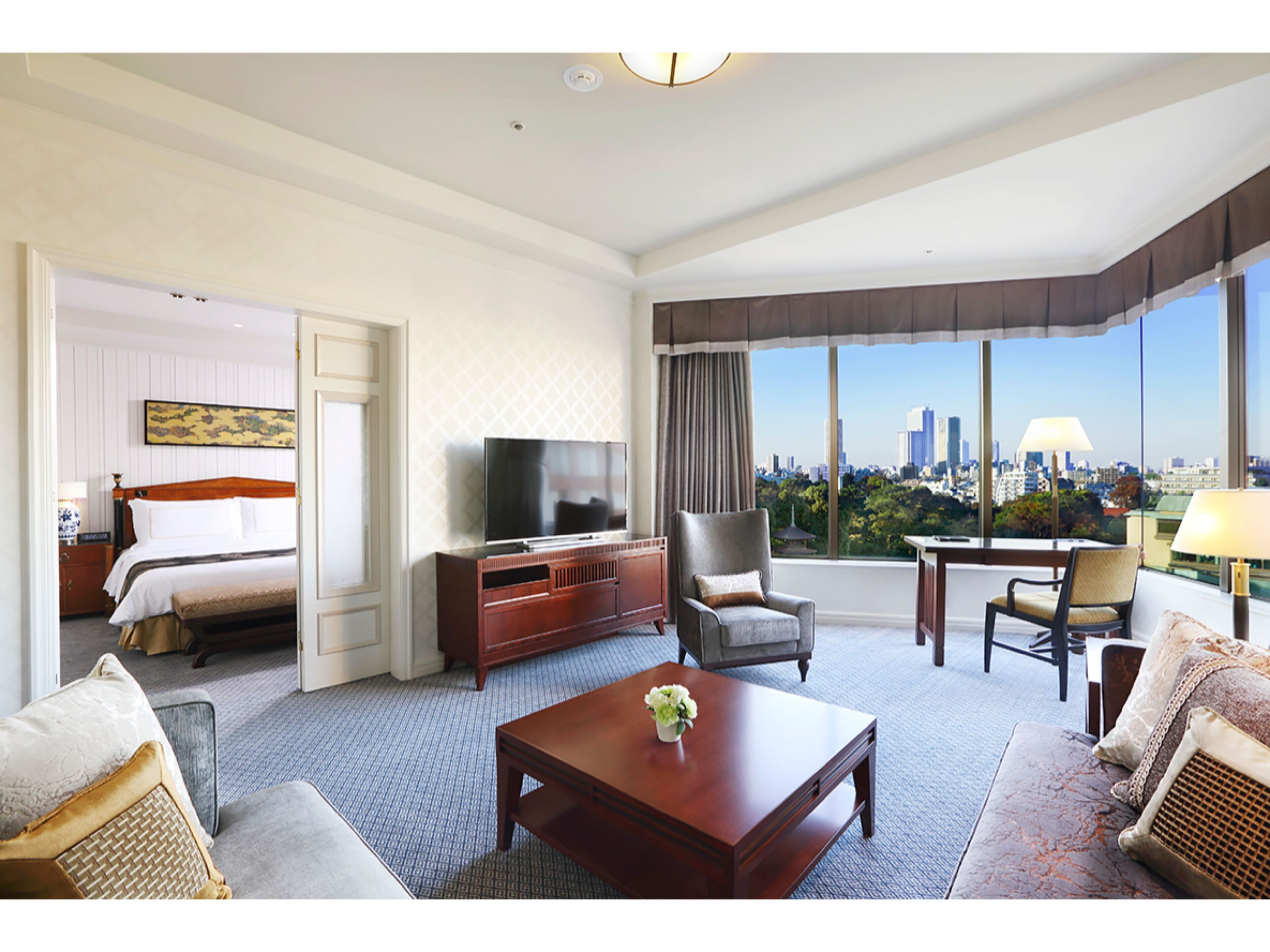 A hotel room with a bed, a TV, a sofa, a coffee table, and a view of a cityscape.