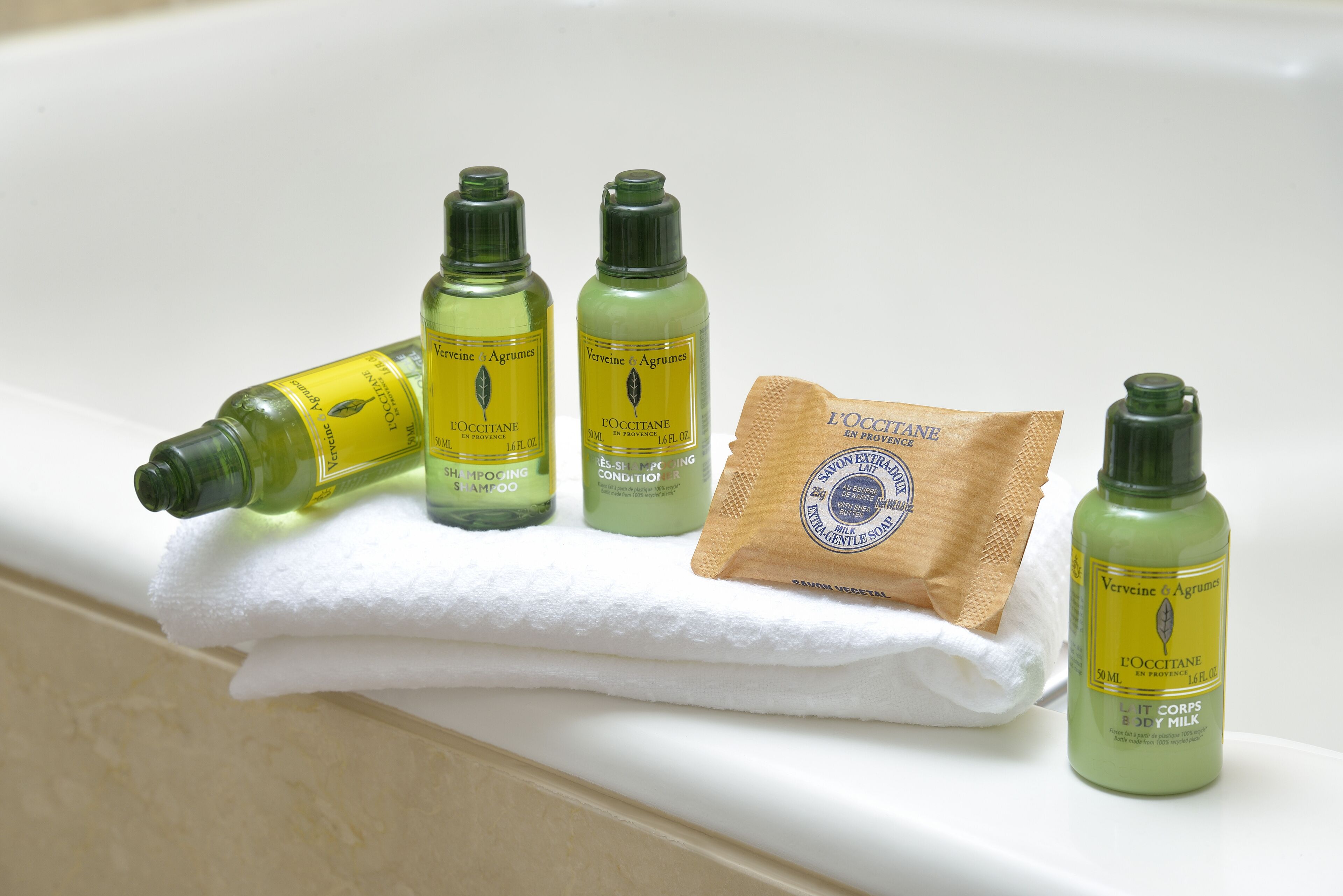 A bathroom with a white bathtub, three bottles of L'OCCITANE body care products, and a brown paper bag.