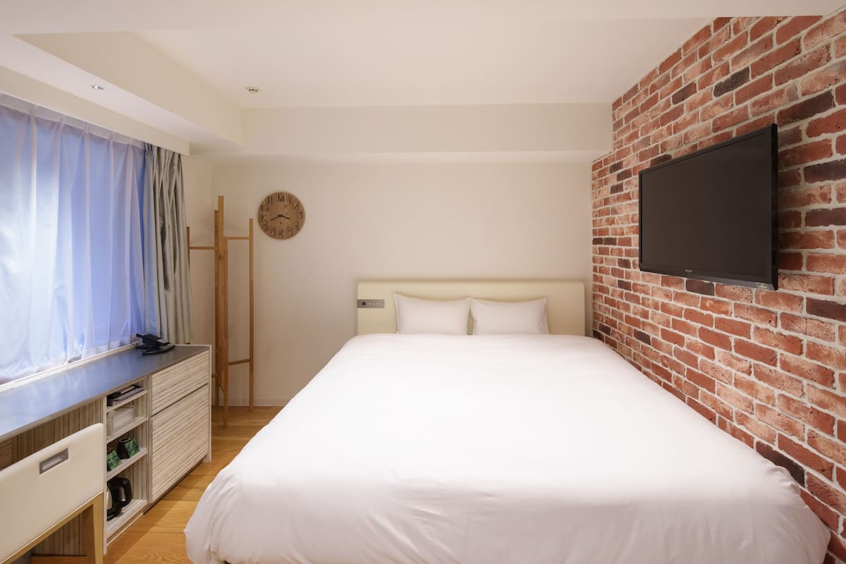 A bedroom with a brick wall, a flat-screen TV, a bed with white bedding, a wooden dresser, and a window with curtains.